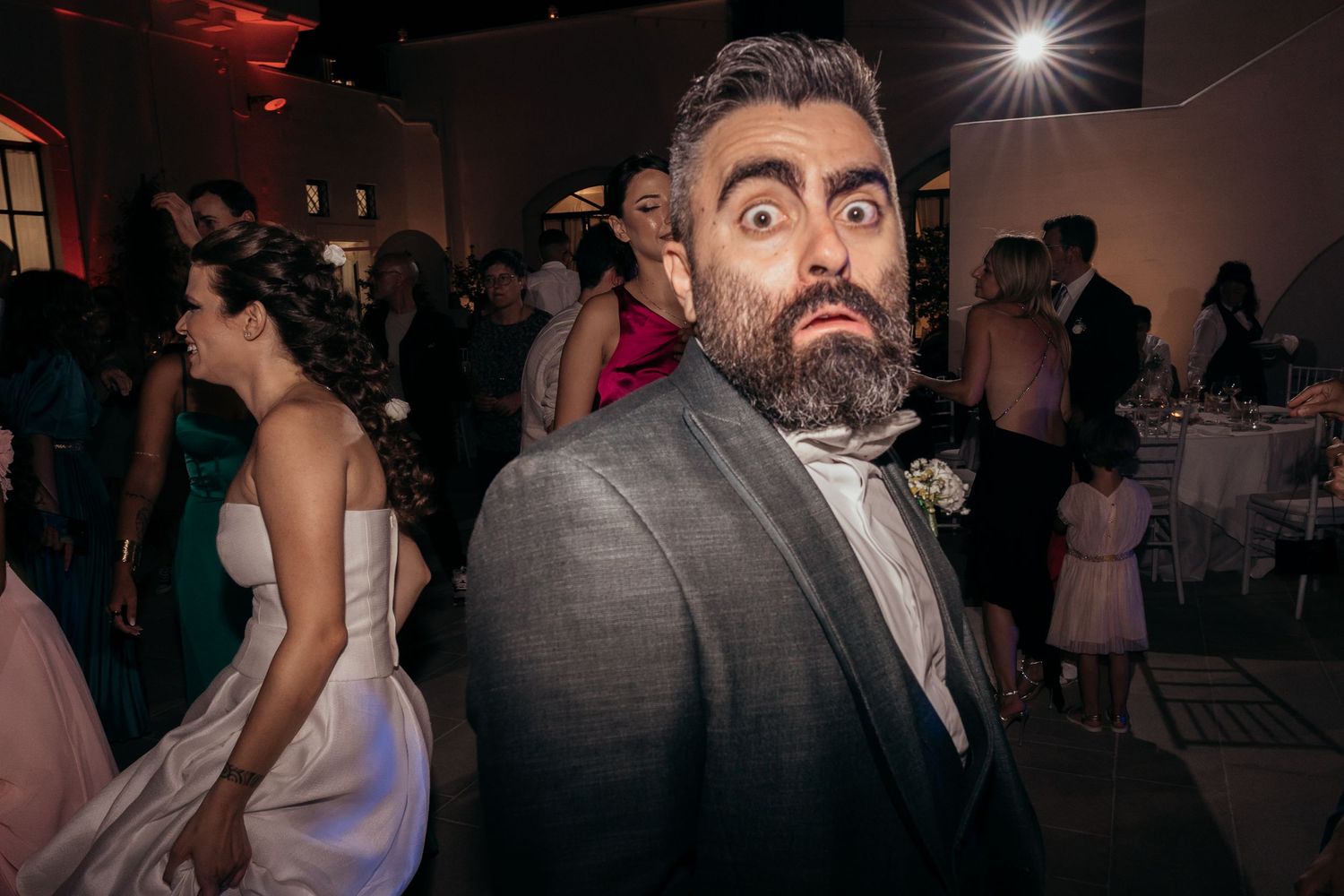 A guest in formal attire makes an expressive face at a dimly lit wedding reception.