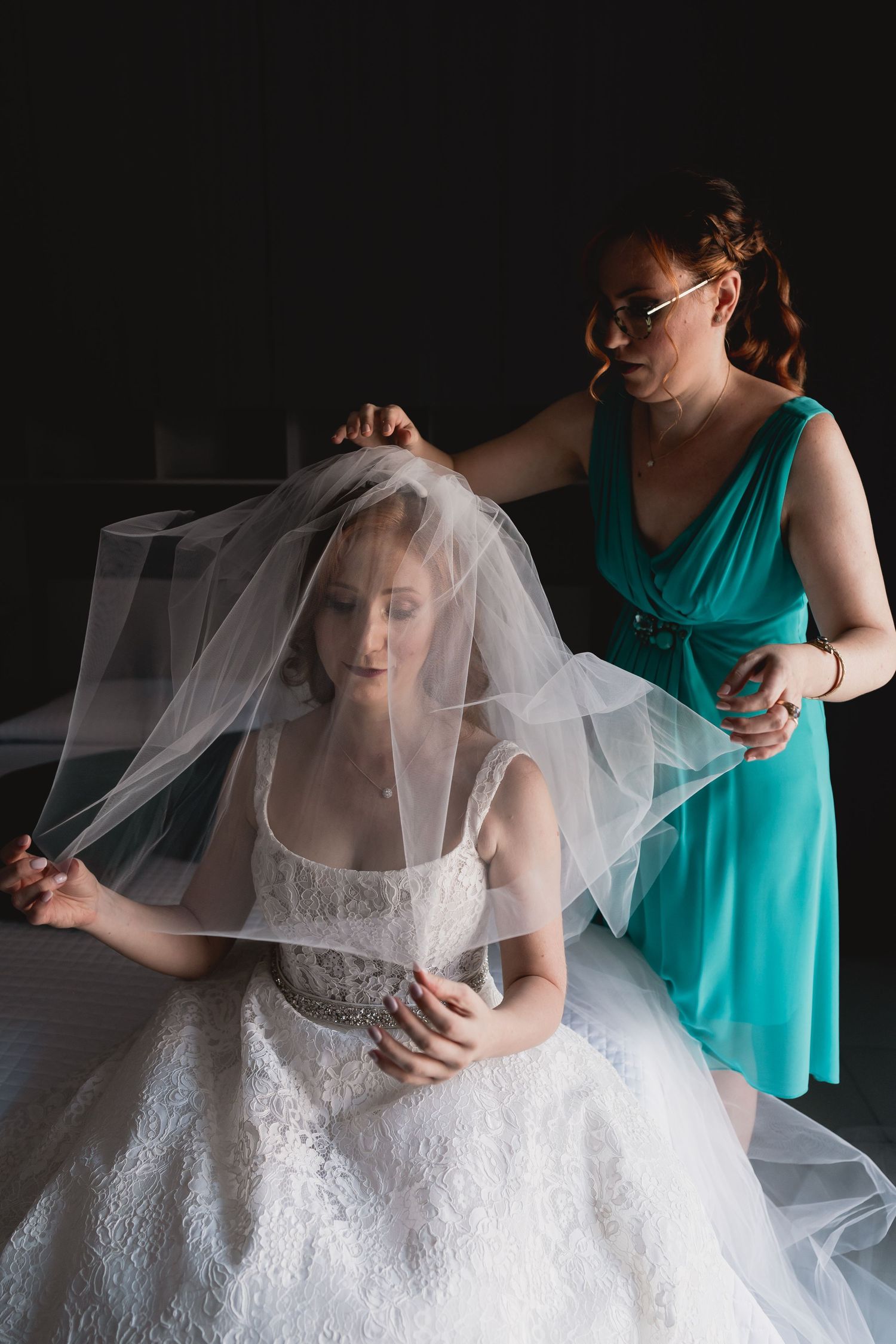 Person in teal dress helps adjust wedding veil in moody lighting.
