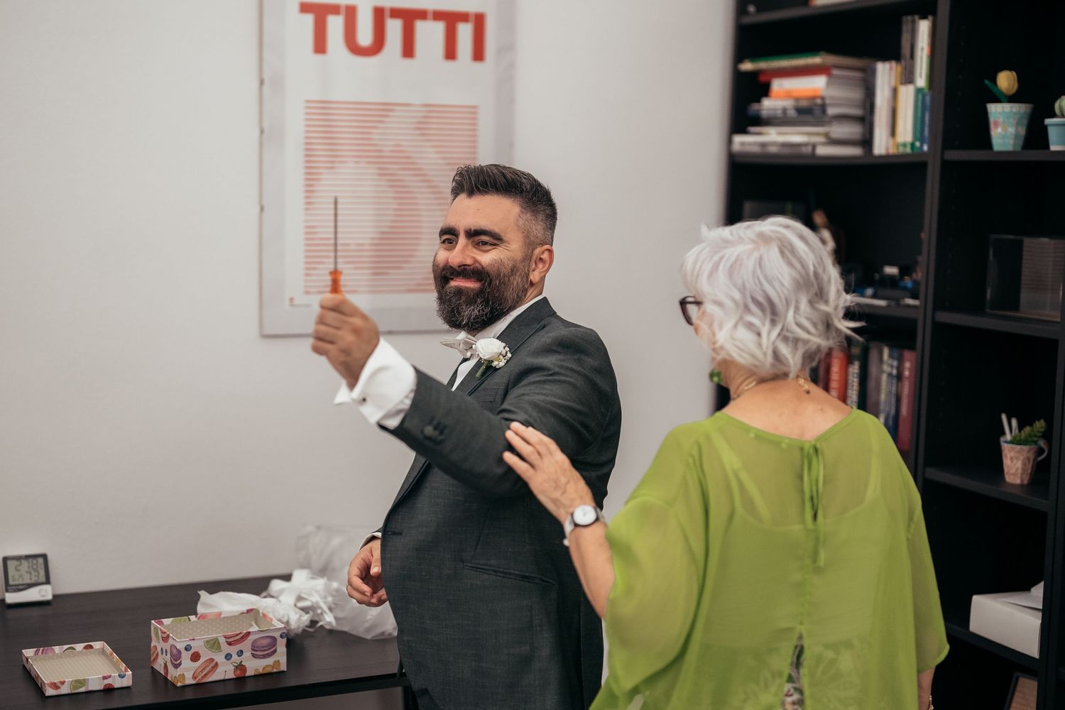 Person in grey suit gesturing while standing next to decorative wall art that says 'TUTTI'.