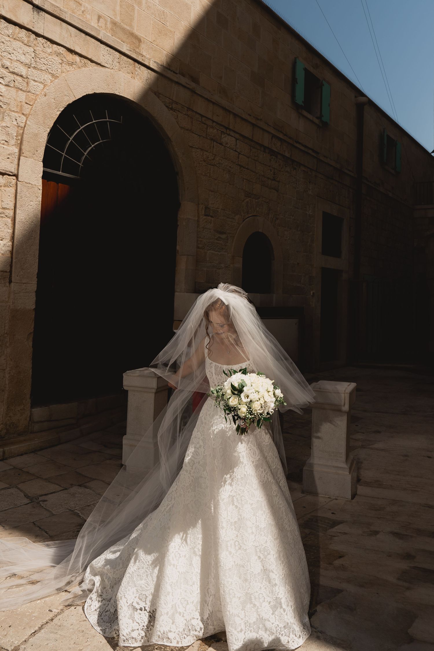 Bride in white lace gown and veil holding bouquet poses in stone archway.