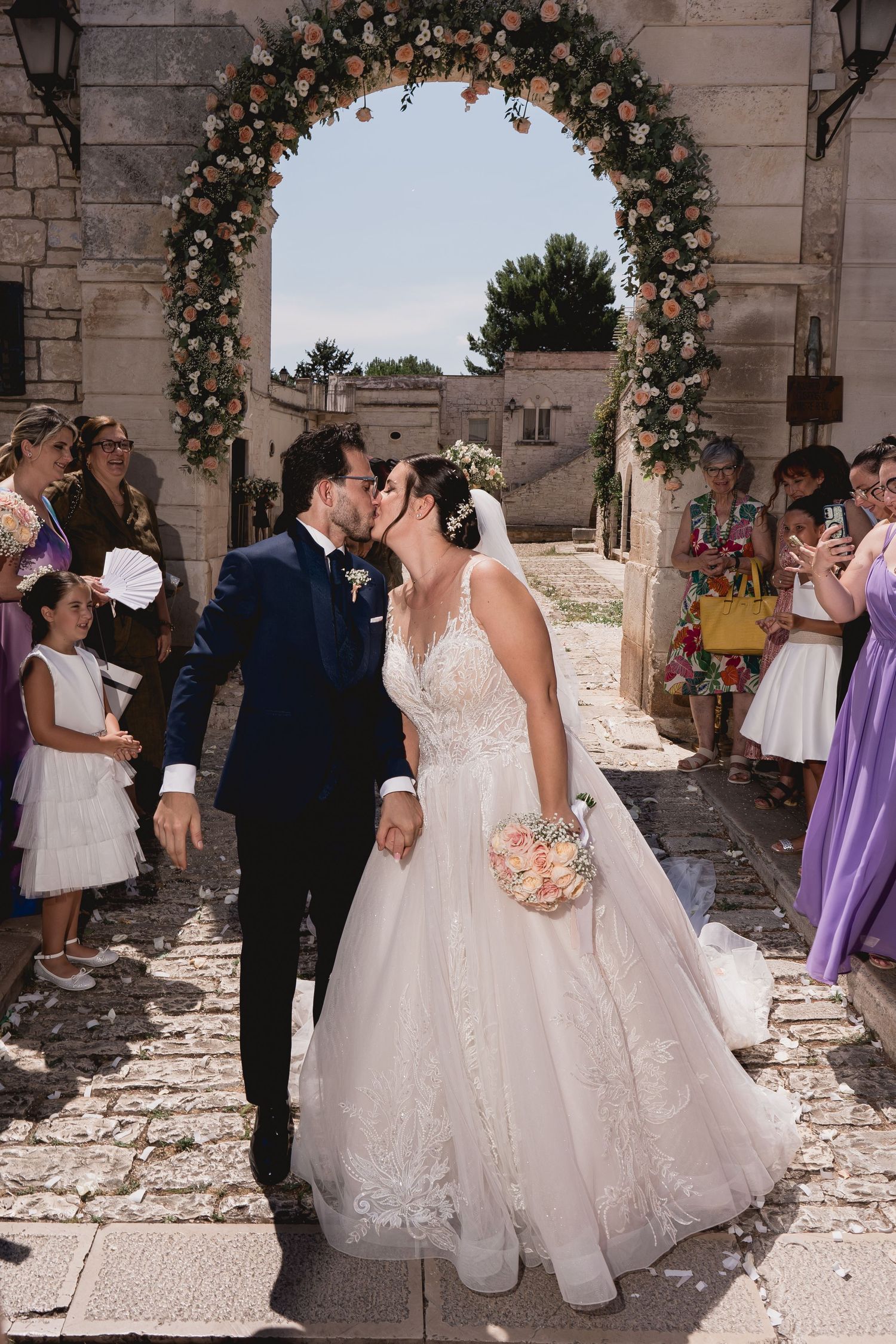Newlyweds share an intimate moment under a floral arch in a historic stone courtyard.