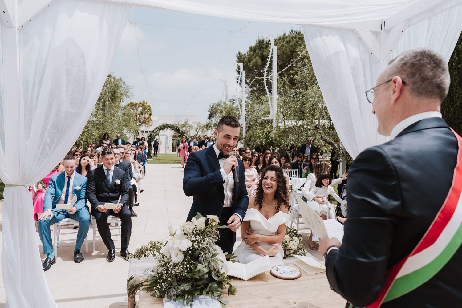 An outdoor wedding ceremony under a white canopy with guests gathered to witness the special moment.