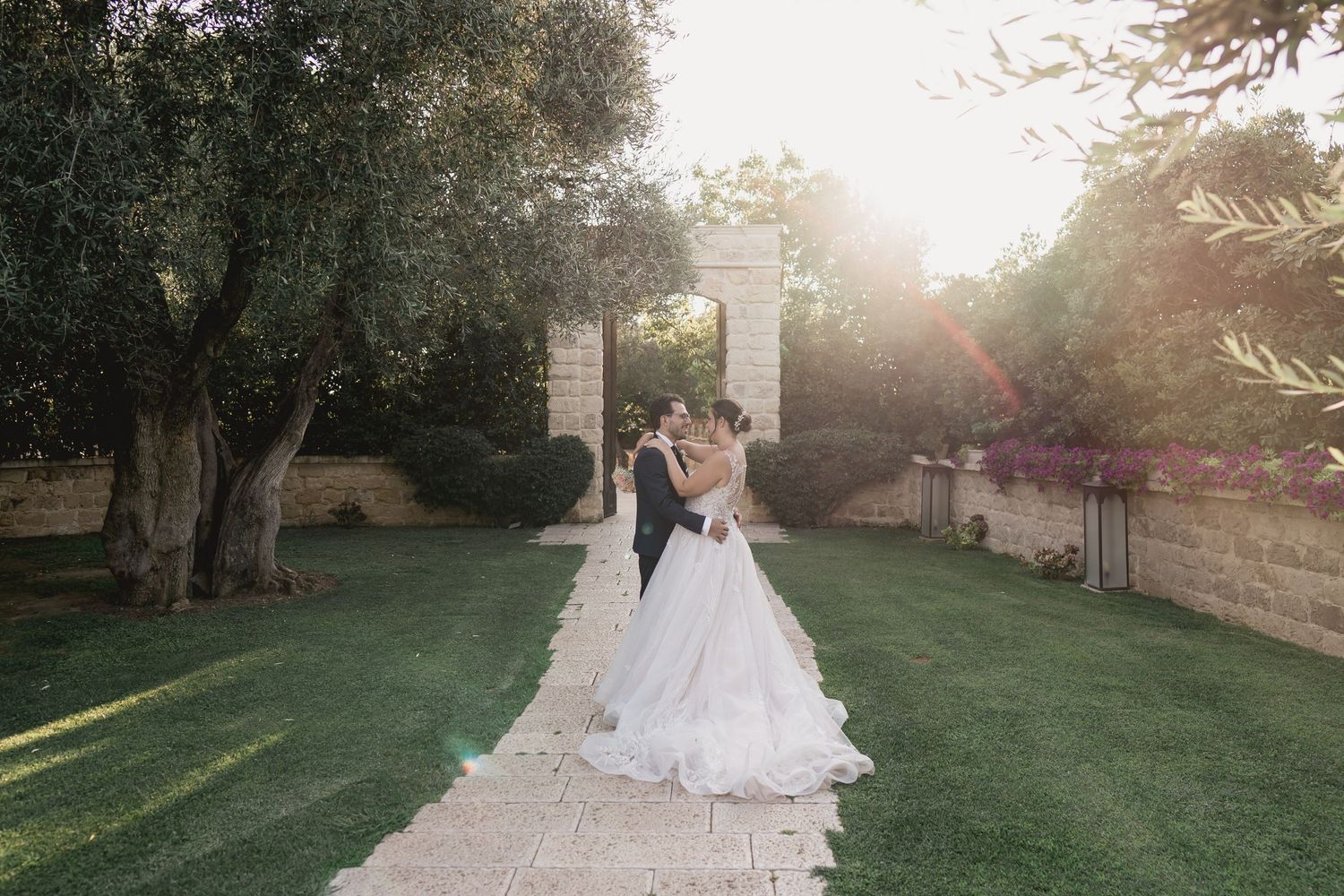 Newlyweds walk down a garden path surrounded by olive trees during golden hour sunset.