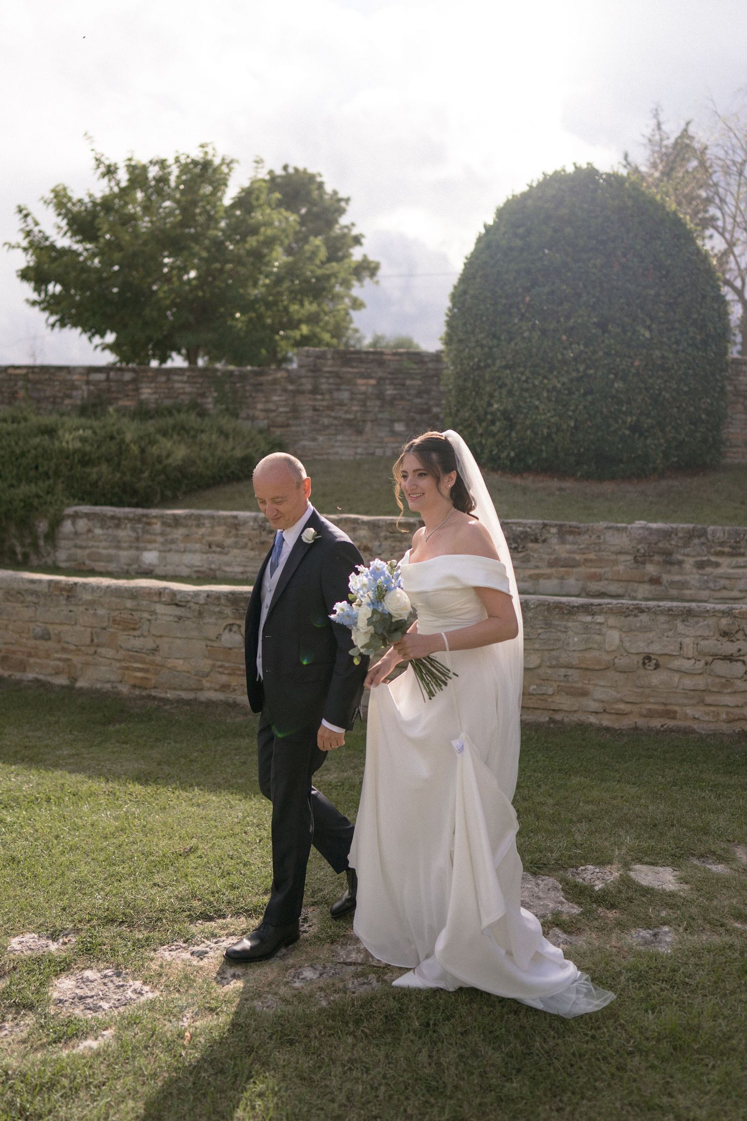 A bride in a flowing white gown walks with someone across a grassy area near a stone wall.