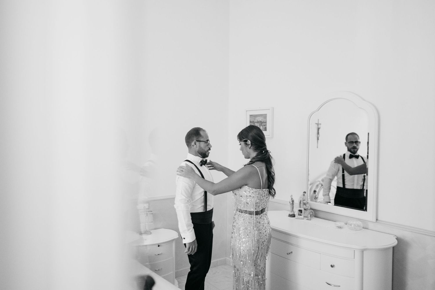 A black and white photo of a couple preparing for their wedding day in front of a vanity mirror.