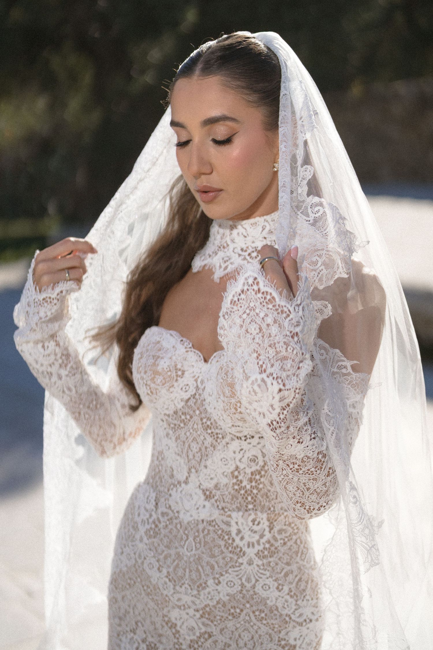 Close-up of a bride in a lace high-neck gown and veil gazing downward with eyes closed as she gently holds the veil.