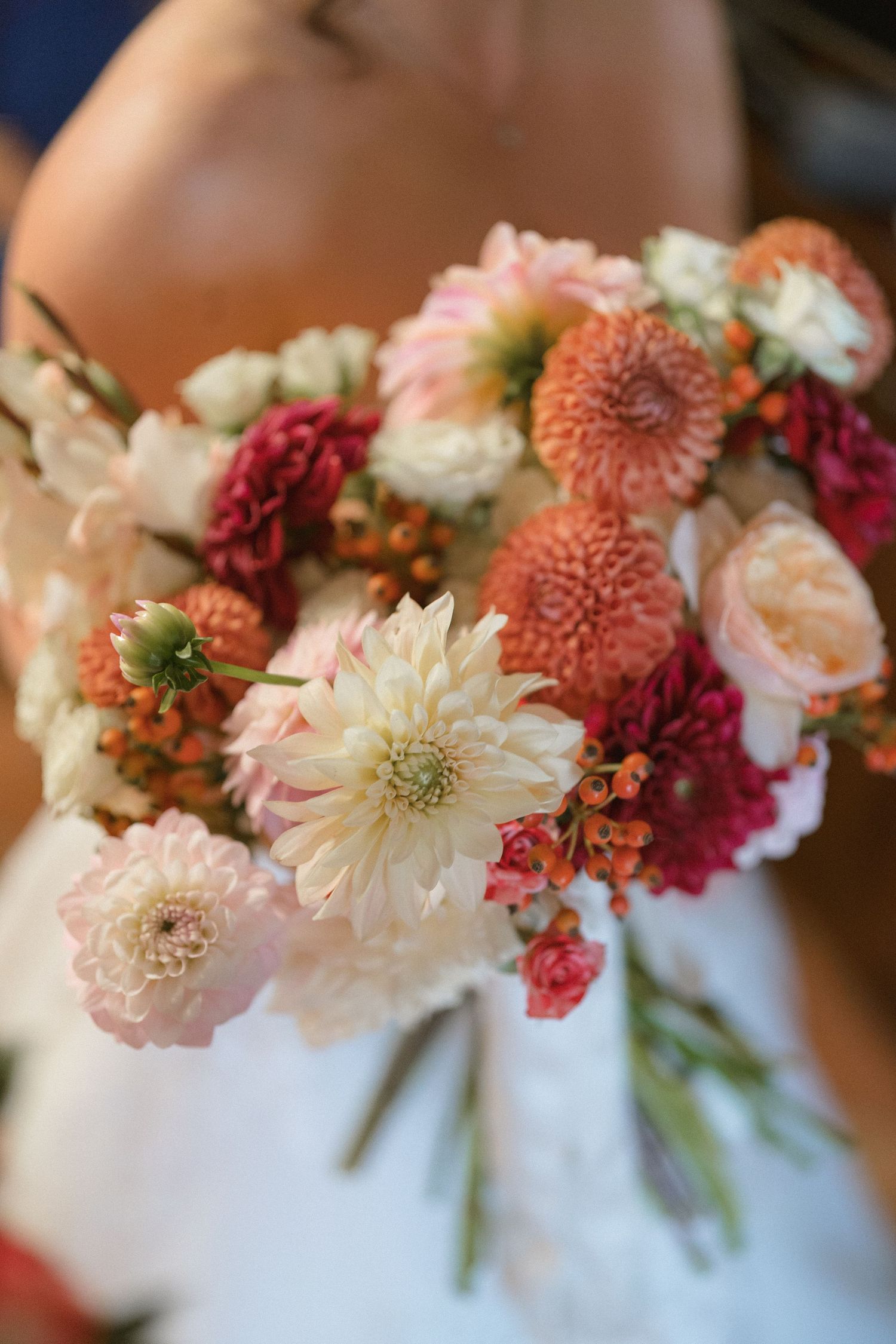 Bridal bouquet featuring coral dahlias, white chrysanthemums, pink roses, and red blooms held by a bride in a white dress.