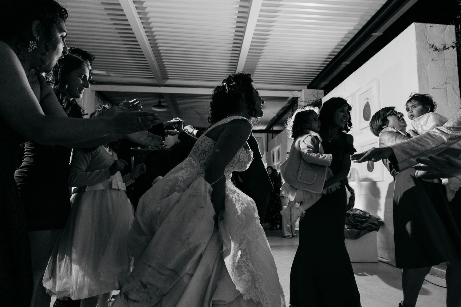 Black and white photo of a bride surrounded by guests dancing at an indoor wedding reception celebration.