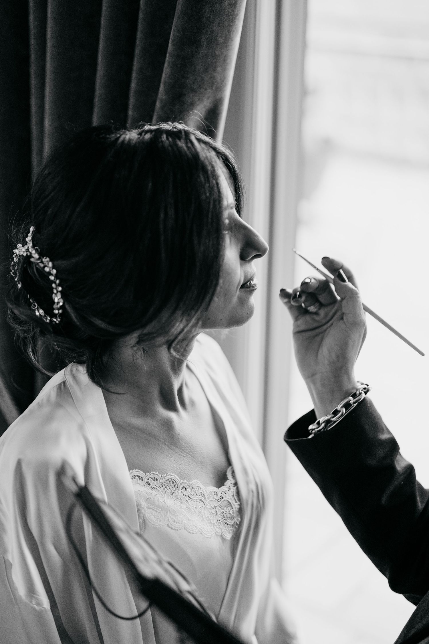 Black and white photo of bride getting makeup applied by artist near window, wearing white robe and floral hair accessory.