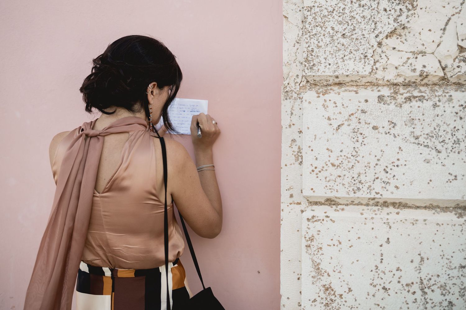 Someone in pink attire leans against a textured wall while holding a phone and camera.