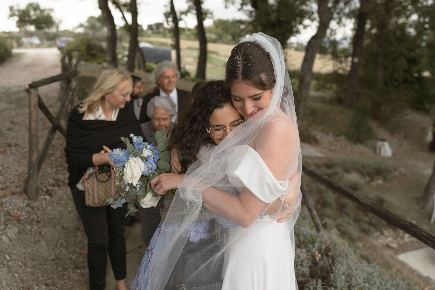 An emotional moment at an outdoor wedding ceremony as attendees embrace while holding a blue bouquet.