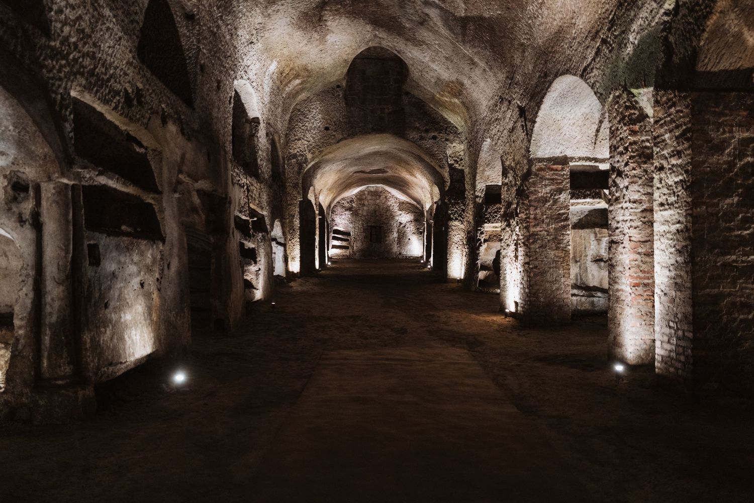 Ancient underground tunnel with arched stone walls and dramatic lighting illuminating the curved pathway stretching into darkness.