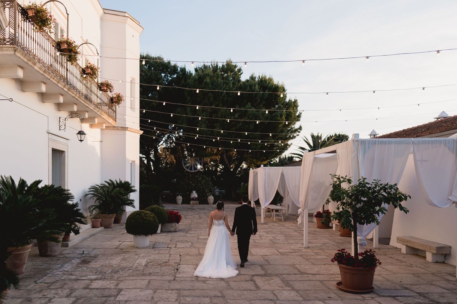 Newlyweds walking through an elegant courtyard with string lights and white fabric drapes.