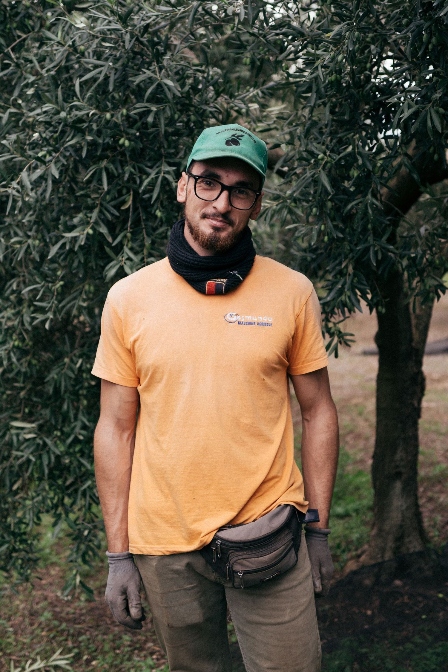 Person wearing orange t-shirt, green cap and headphones stands in front of leafy trees in casual outdoor setting.