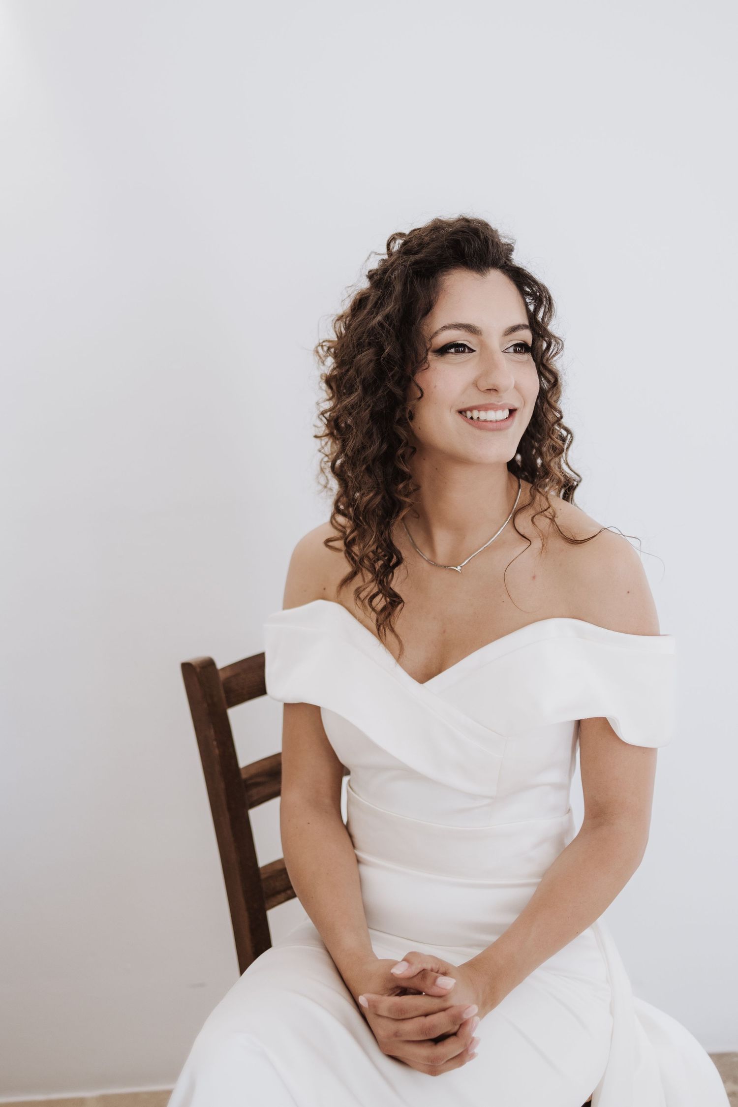 A bride in an elegant off-shoulder white wedding dress sits on a wooden chair with natural curly hair.