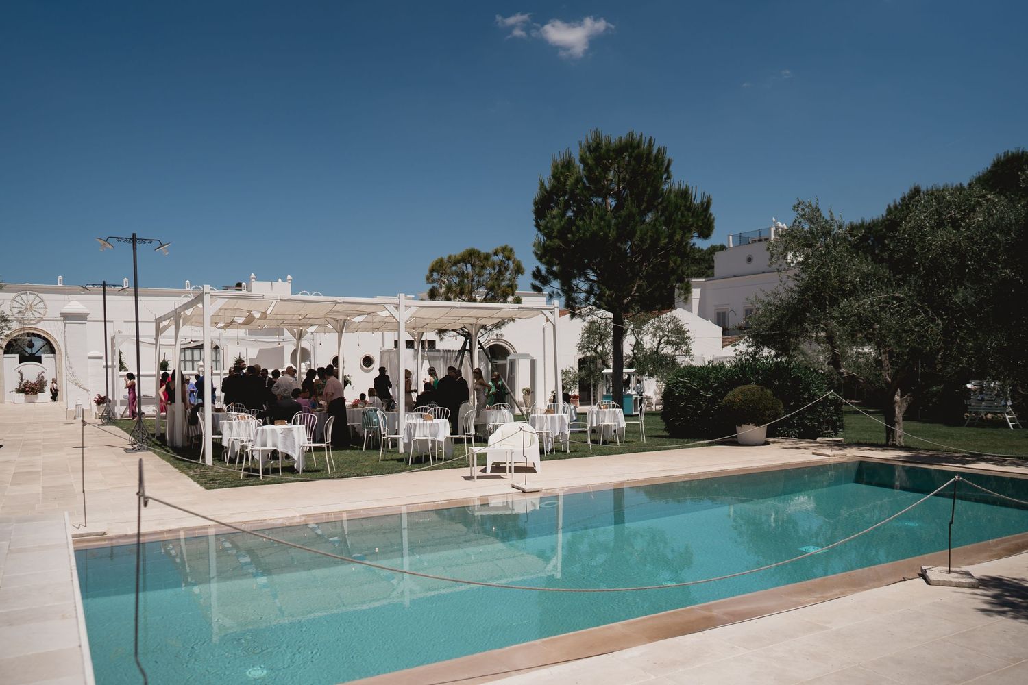 White Mediterranean-style venue with swimming pool and outdoor seating area under bright blue sky.