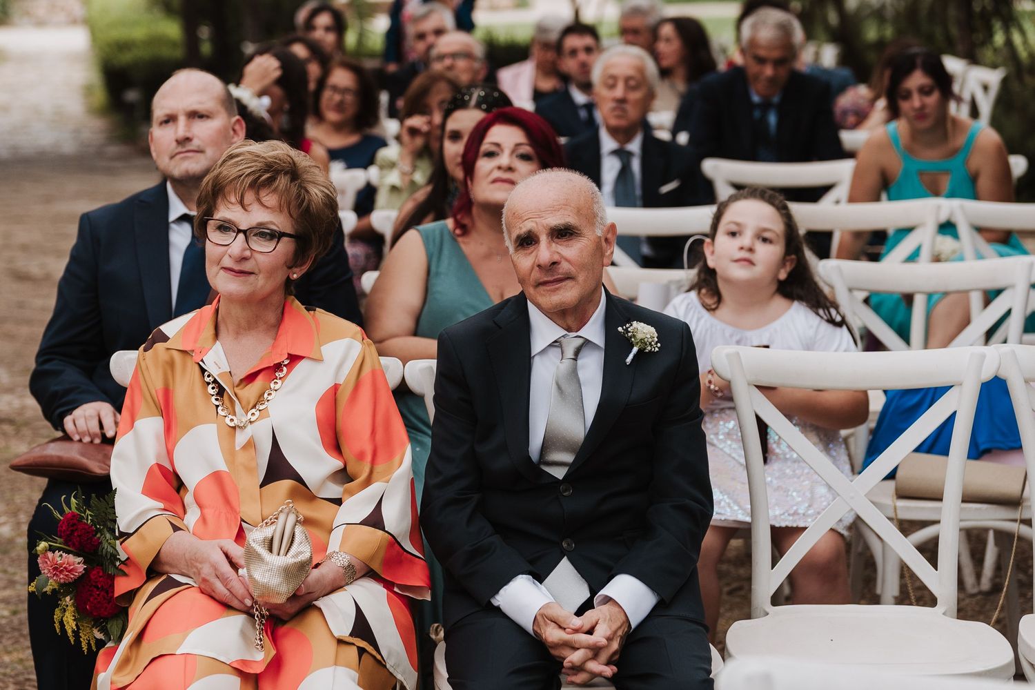 Wedding guests seated in white chairs watch outdoor ceremony attentively.