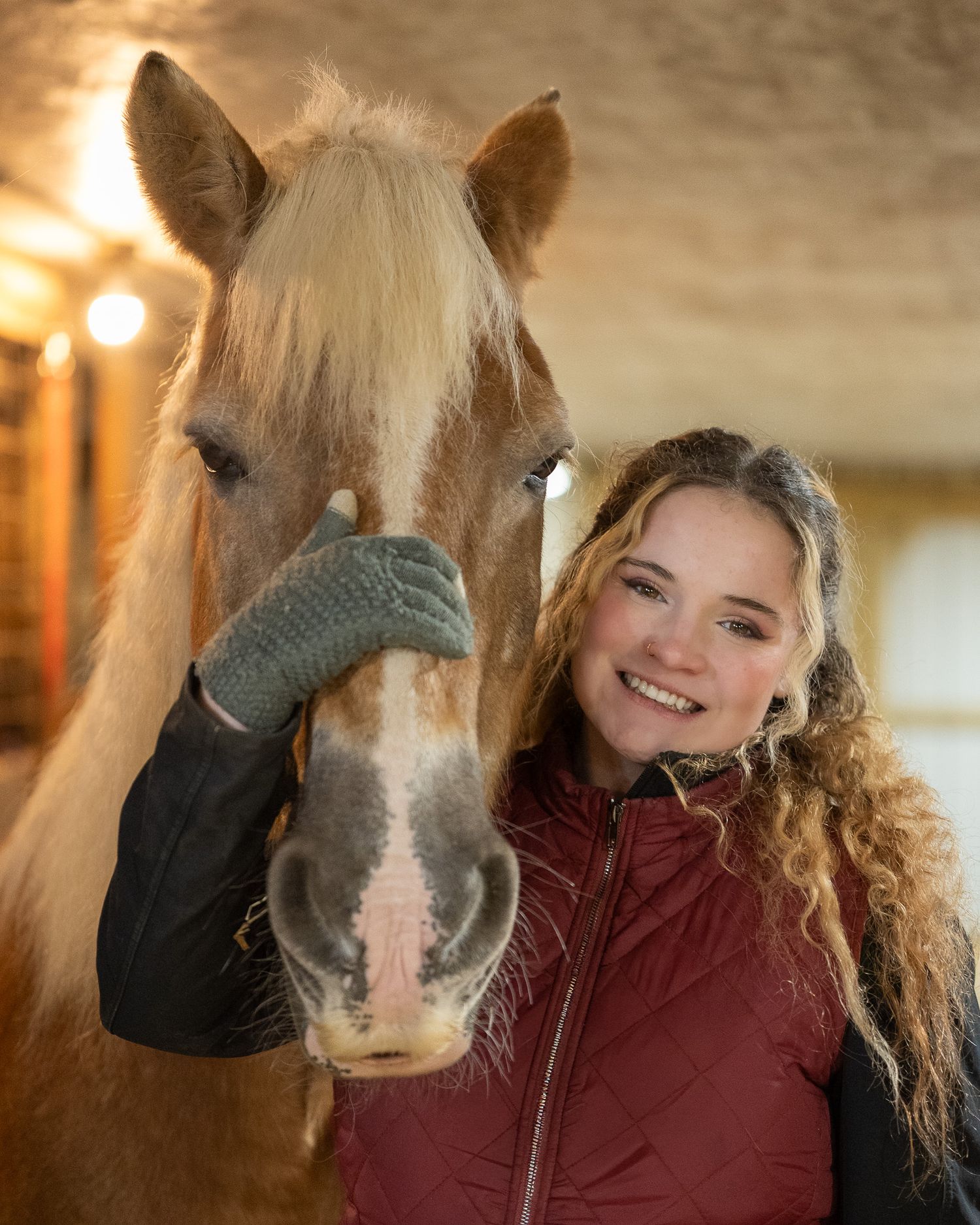 A person in a burgundy jacket poses playfully with a brown horse in a stable.