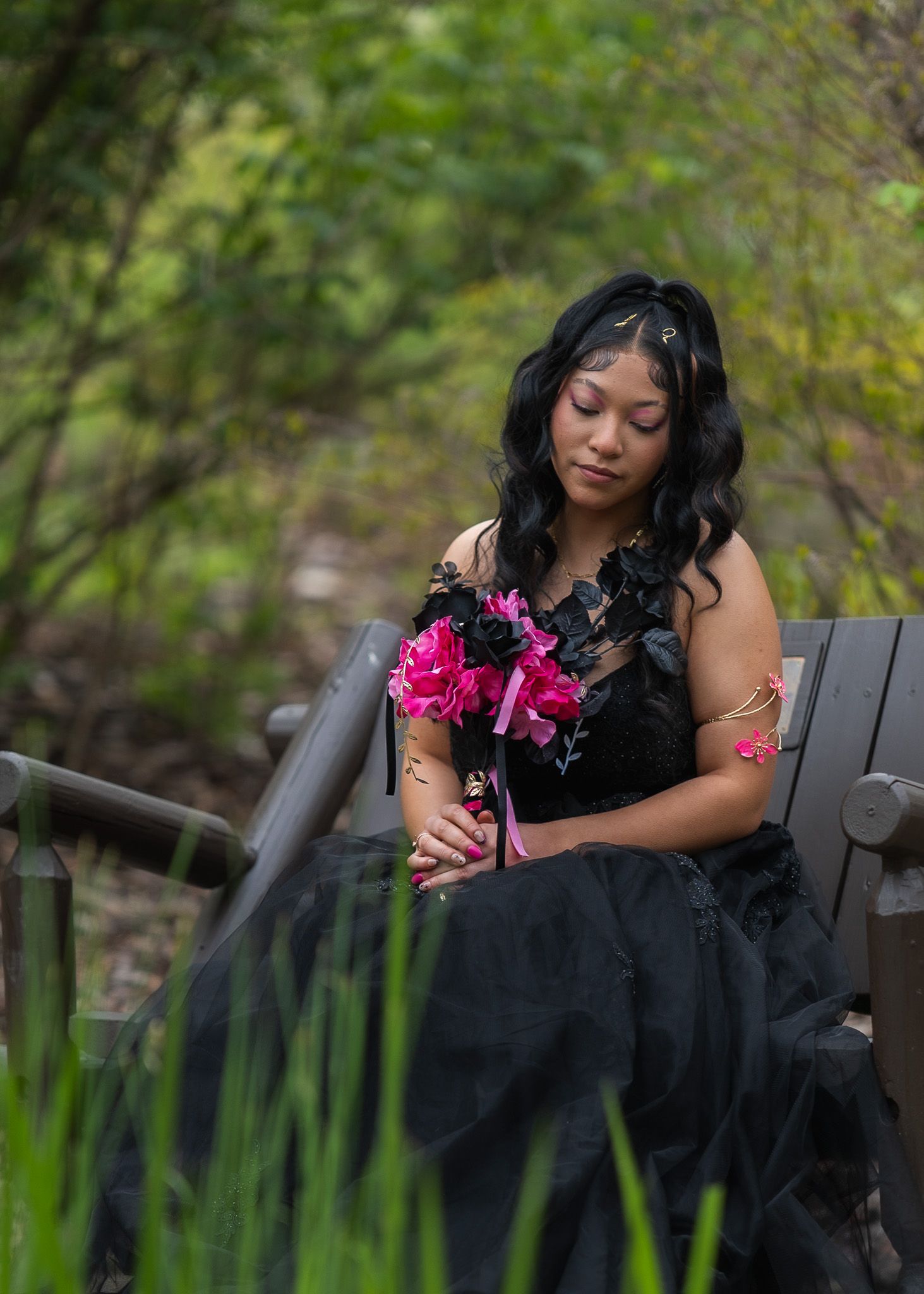 Young woman in black gown holding pink flowers, seated on bench surrounded by lush green foliage.