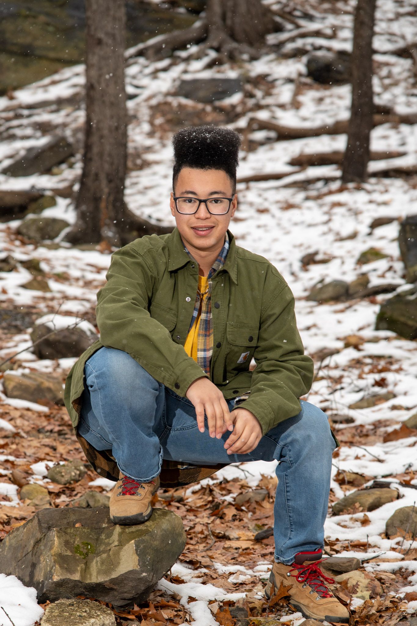 A person in a green jacket and jeans sits on rocky ground with patches of snow in a forest setting.