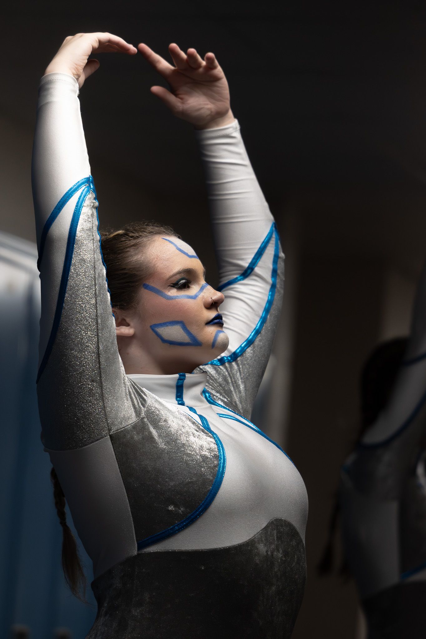 Young performer in gray and blue costume with artistic face paint raises arms gracefully during a color guard routine.