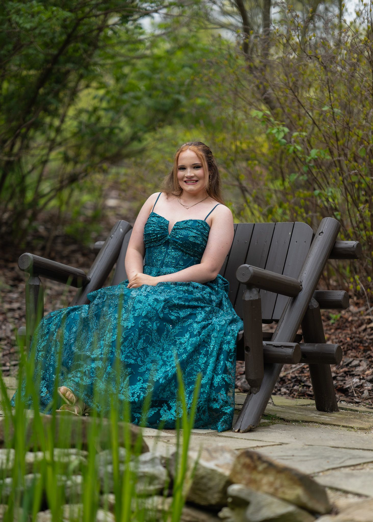A young woman in a teal lace ball gown sits gracefully on a dark wooden bench surrounded by lush garden greenery.