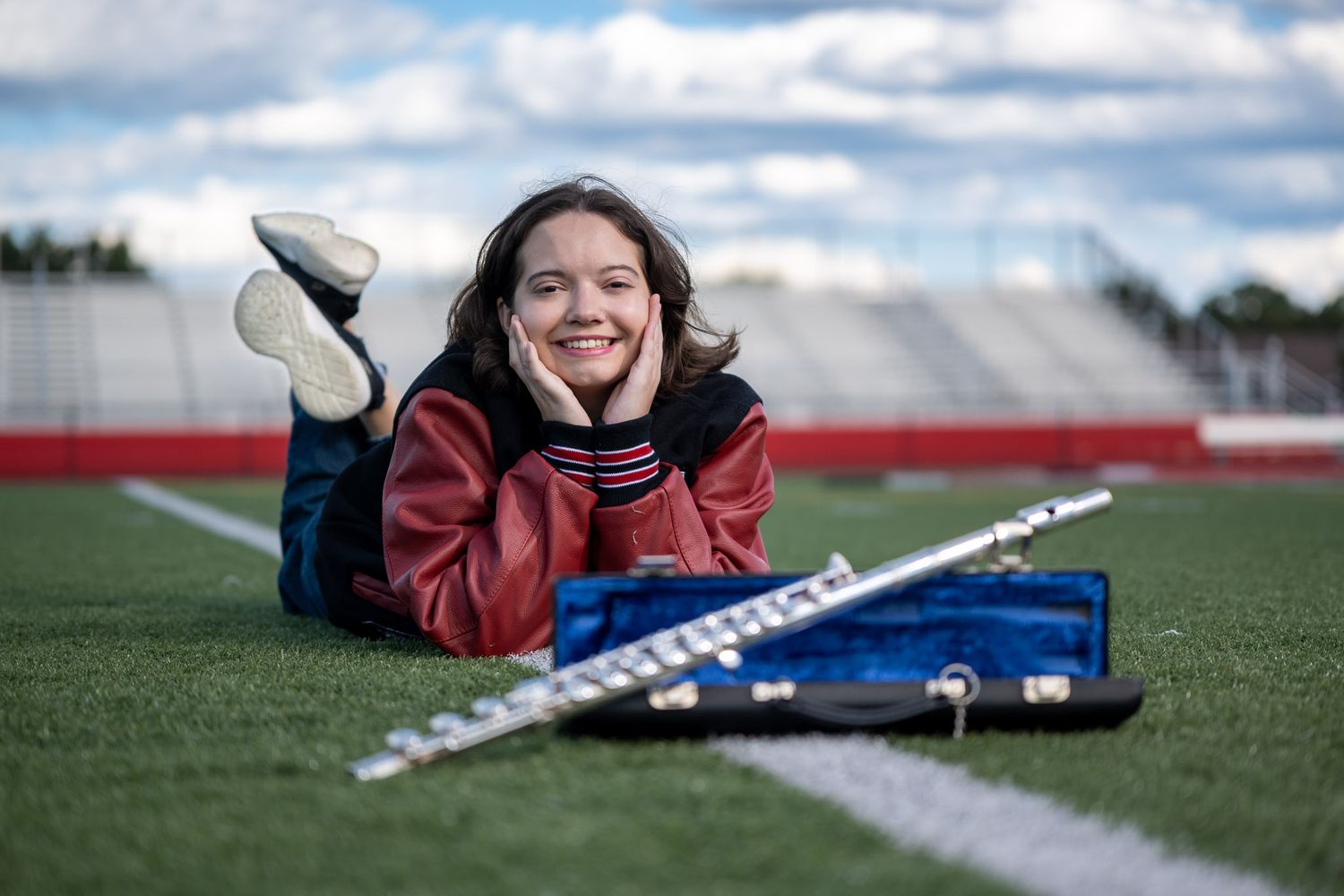 A smiling girl lies on green turf holding her face, with a flute and case in front of her at a stadium.