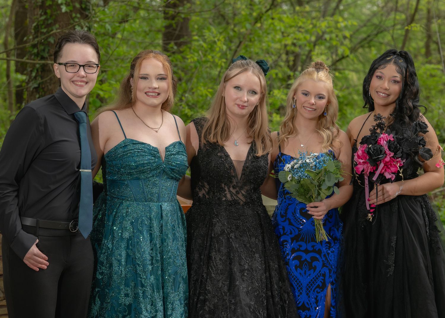 Five teenagers in formal prom attire pose together outdoors among trees, wearing elegant gowns and suits.