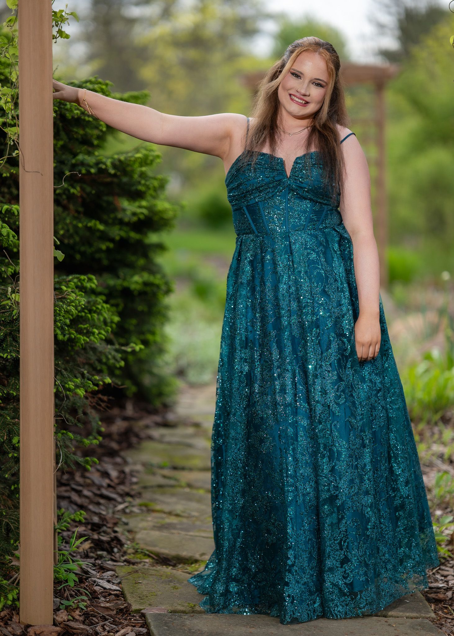 Young woman in teal sequin gown poses gracefully in garden, wearing elegant floor-length formal dress for prom.