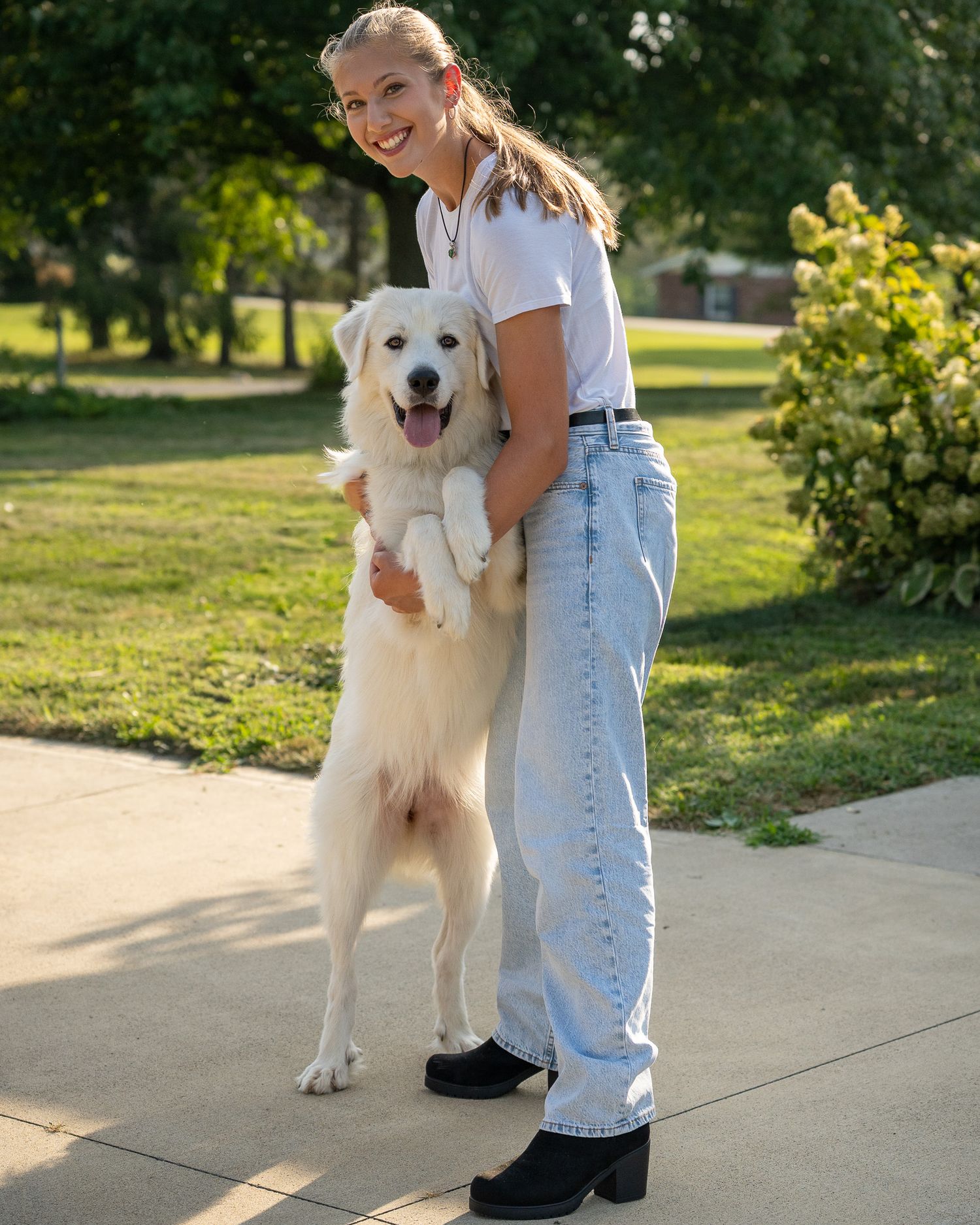 A person in denim overalls stands with a friendly white dog on a sunny pathway.