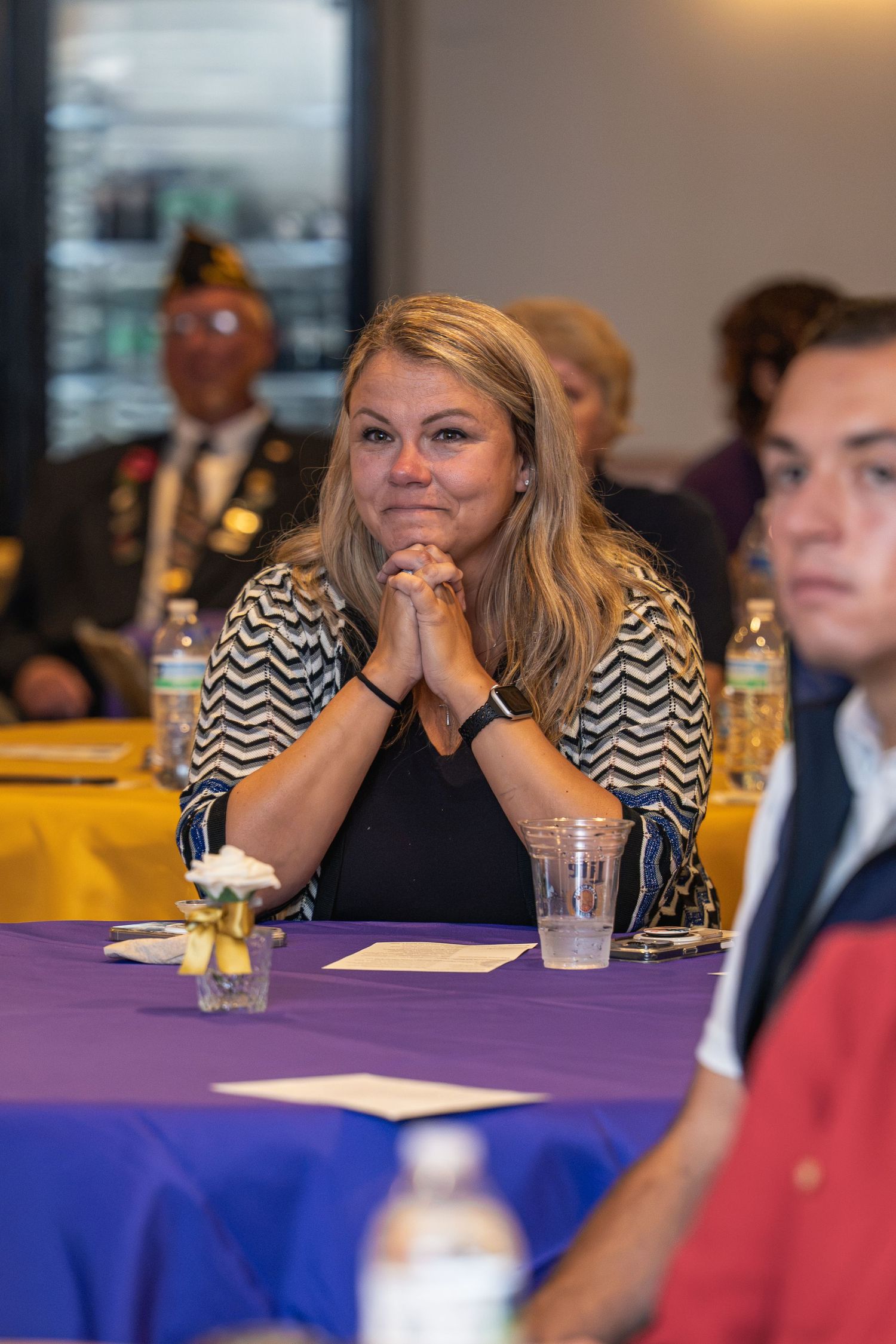 Emotional blonde woman sits at a purple-draped banquet table with hands clasped during a formal event.
