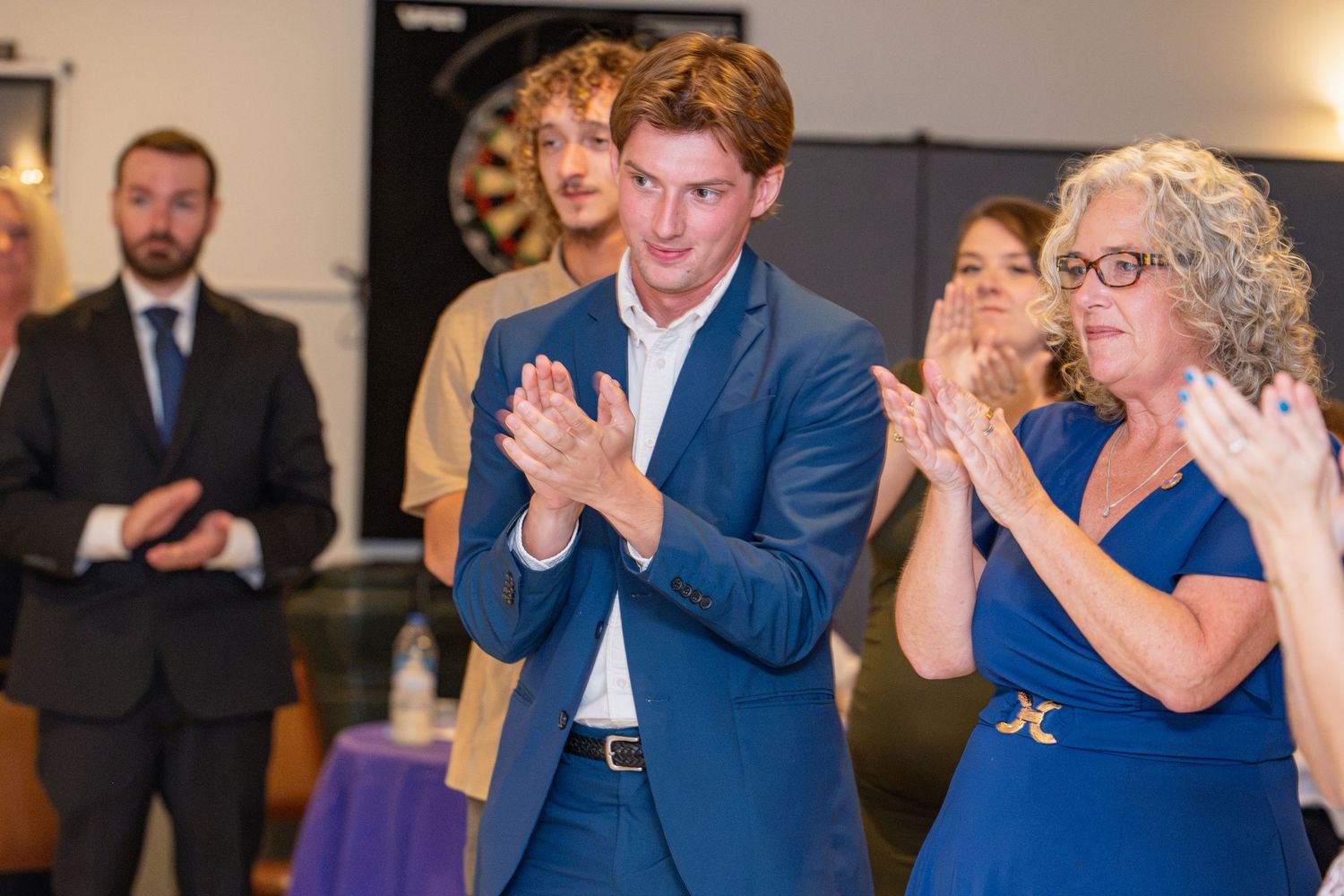 Young man in blue suit clapping at formal event with woman beside him and guests in background.