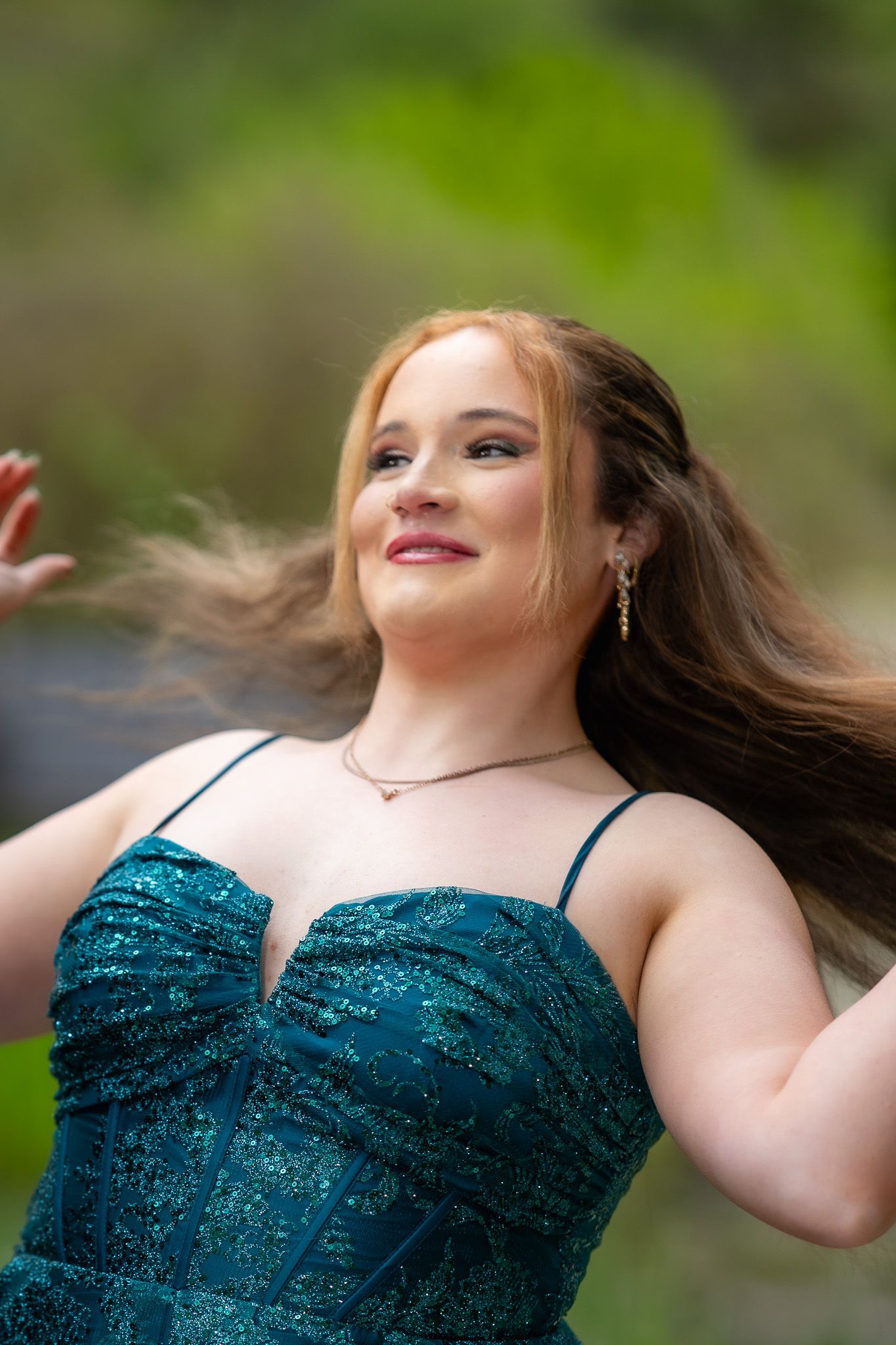 Young woman in teal sequin gown with flowing red hair, smiling outdoors with green foliage background.
