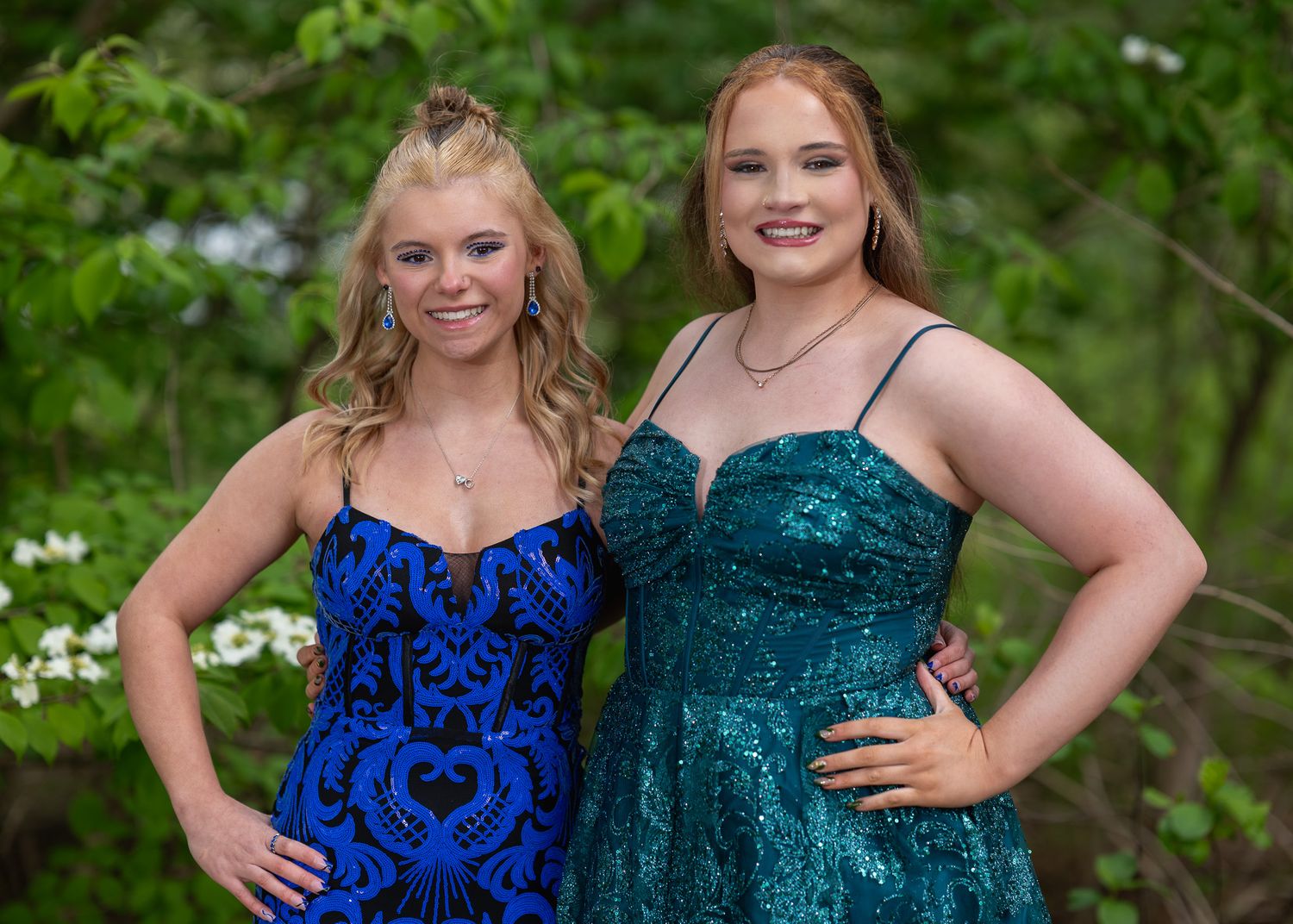 Two young women in sparkly prom dresses, blue and teal, smiling outdoors among green trees.