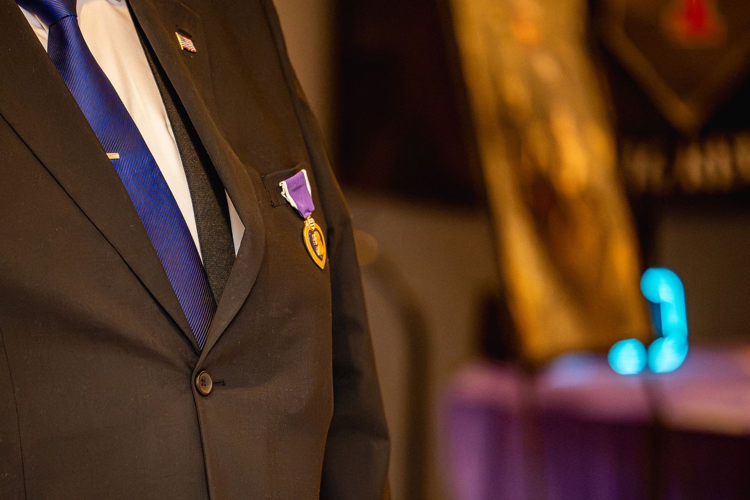 Man in dark suit wearing a Purple Heart military medal with blue tie at a formal ceremony.