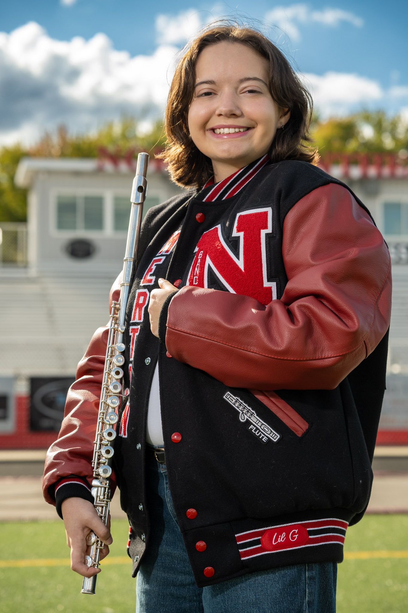 Band member in letterman jacket holding a flute instrument outdoors.