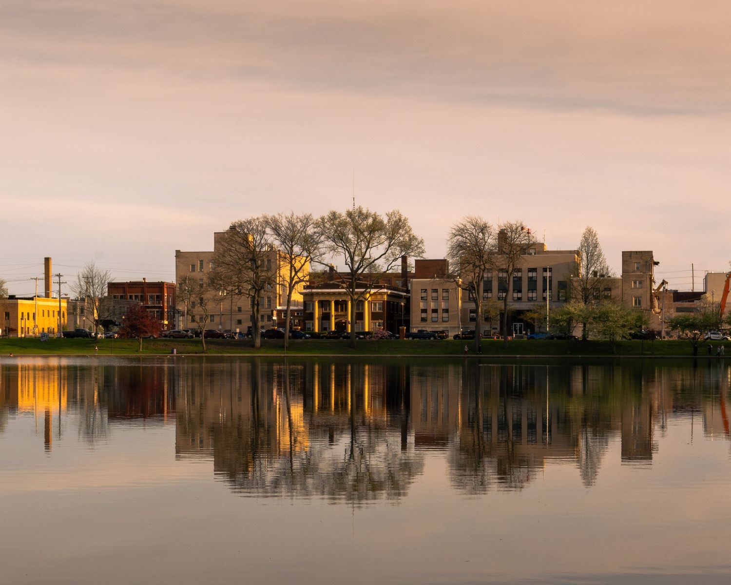 Panoramic view of buildings reflected in calm lake waters during sunset with golden light illuminating the scene. Barberton, OH