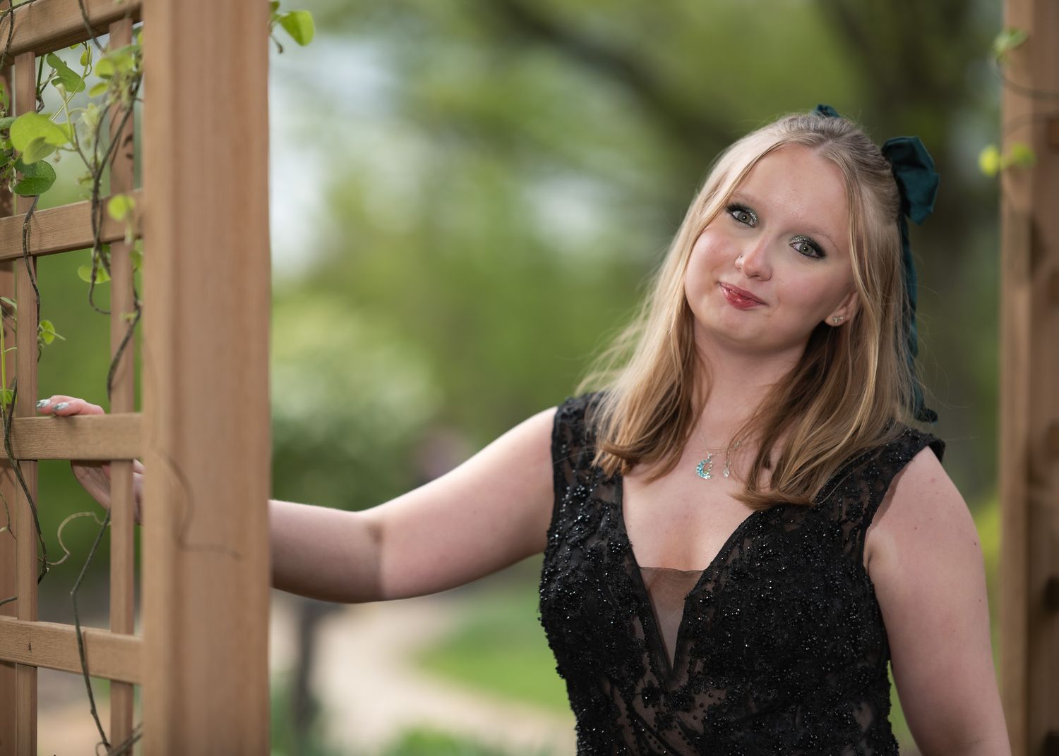 Young blonde woman in black lace dress posing by wooden garden trellis with green vines outdoors.