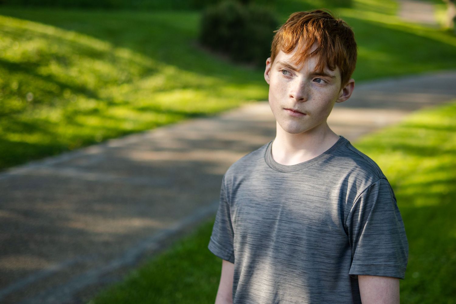 Portrait of youth in grey t-shirt on path with sunlit green background.