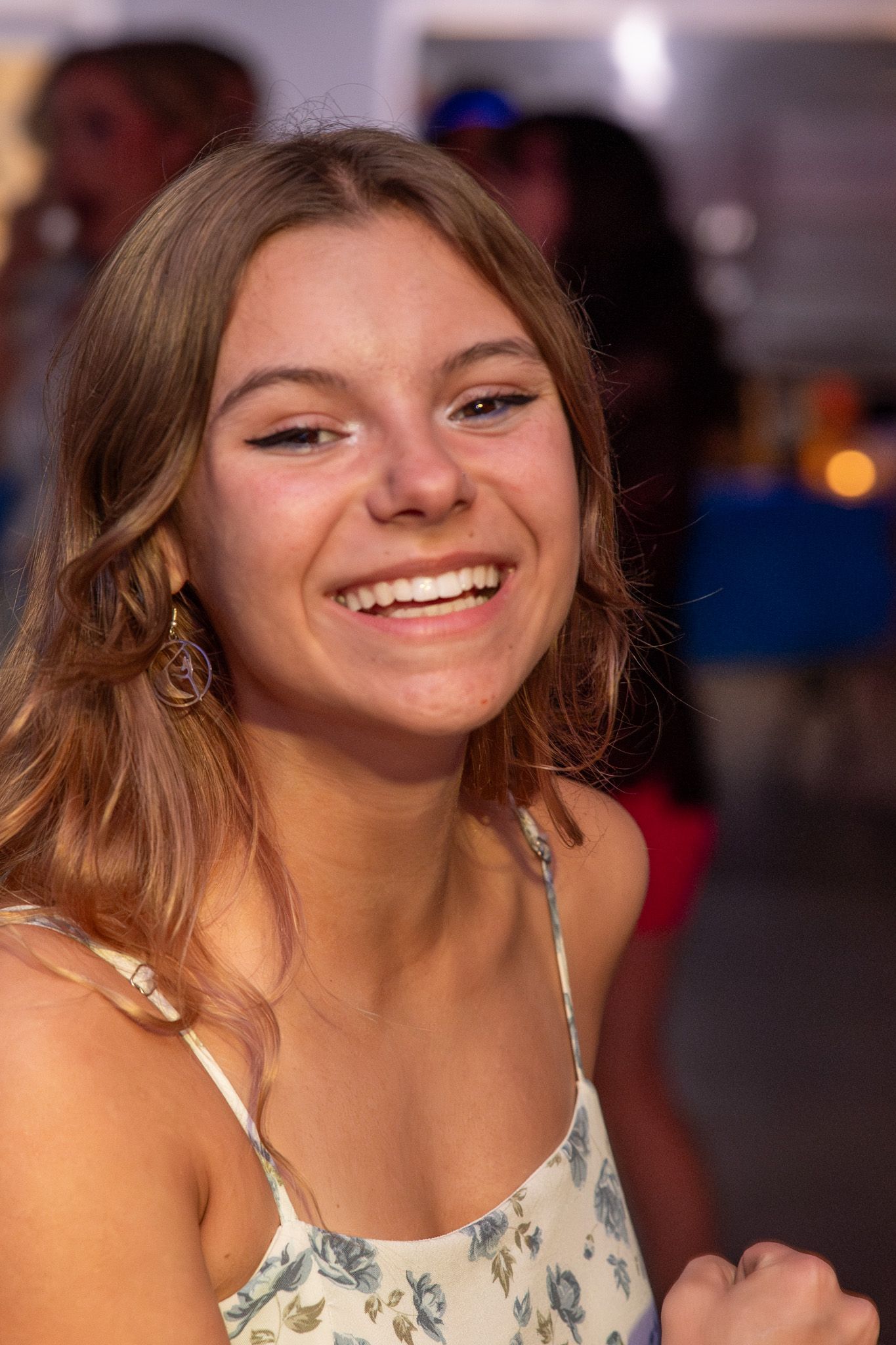 Smiling young woman with brown hair wearing floral dress and gold earrings at a social gathering.