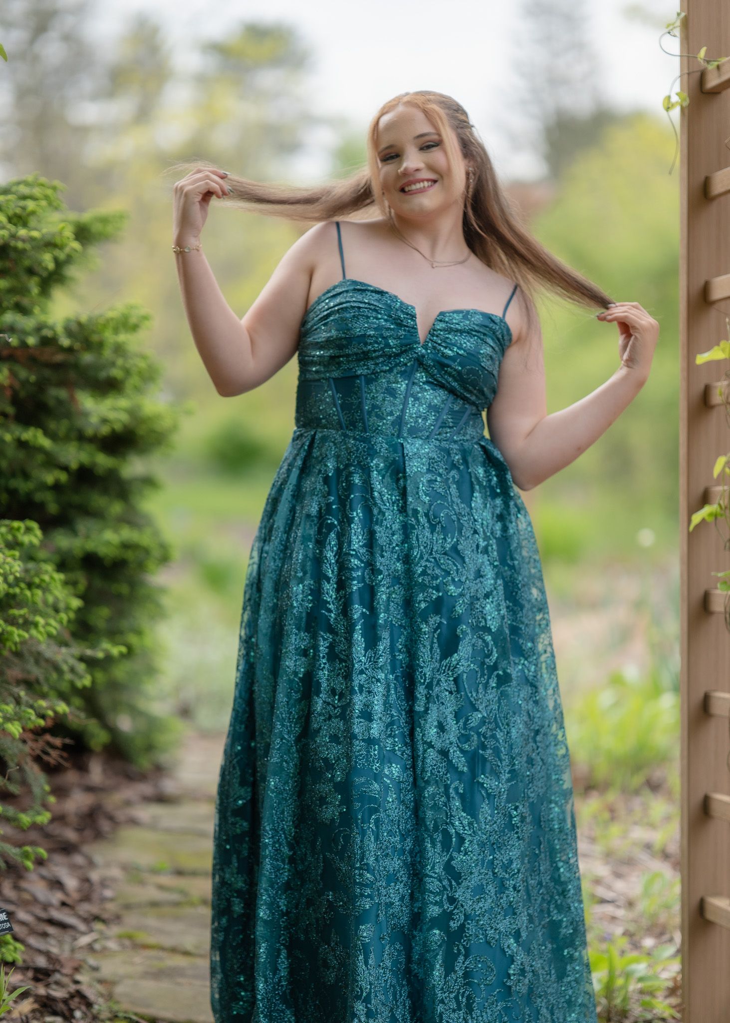 Woman in teal lace A-line gown posing outdoors in a garden setting, smiling and holding her hair.