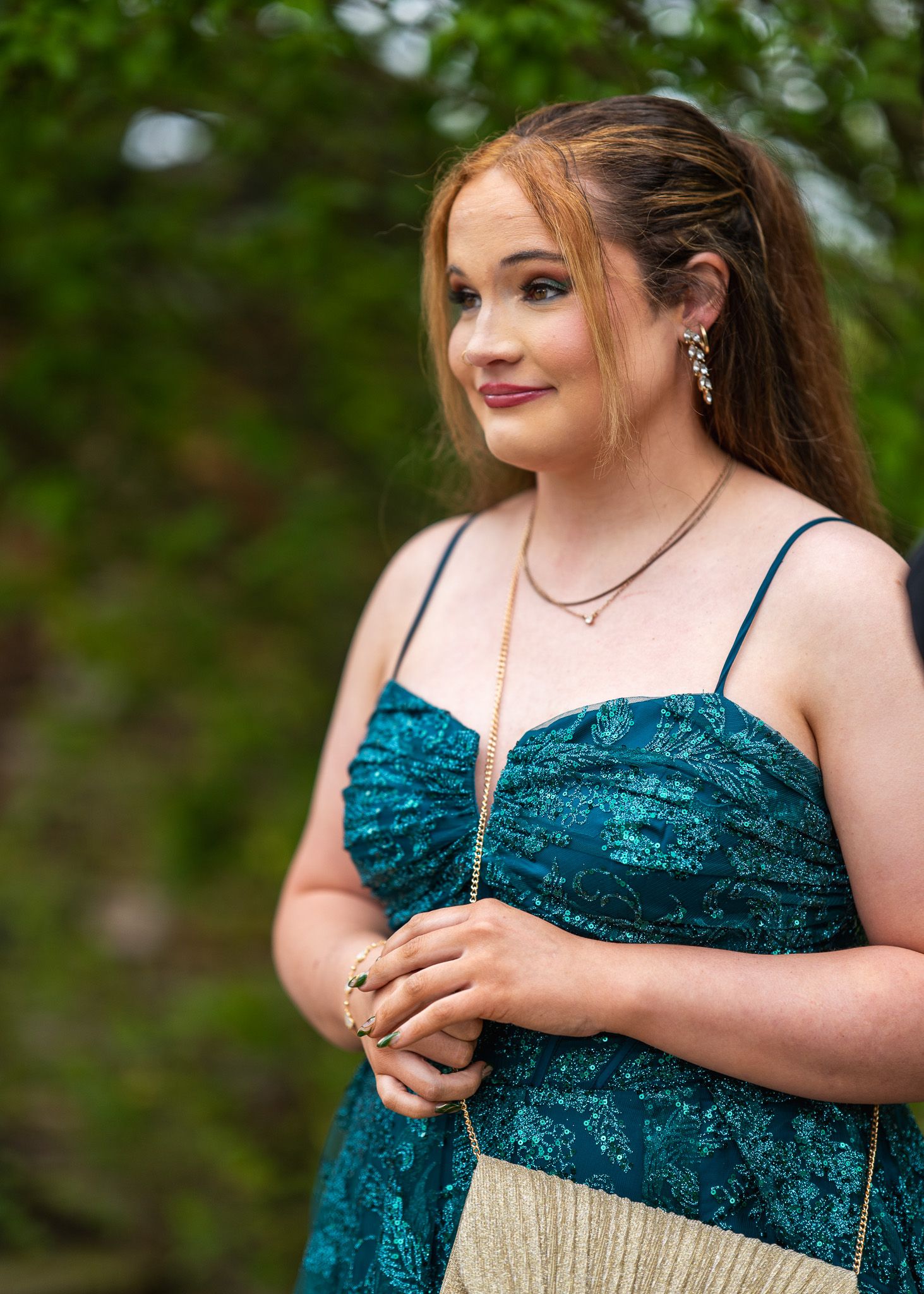 Young woman in teal sequin dress with gold accessories poses outdoors near lush green trees.