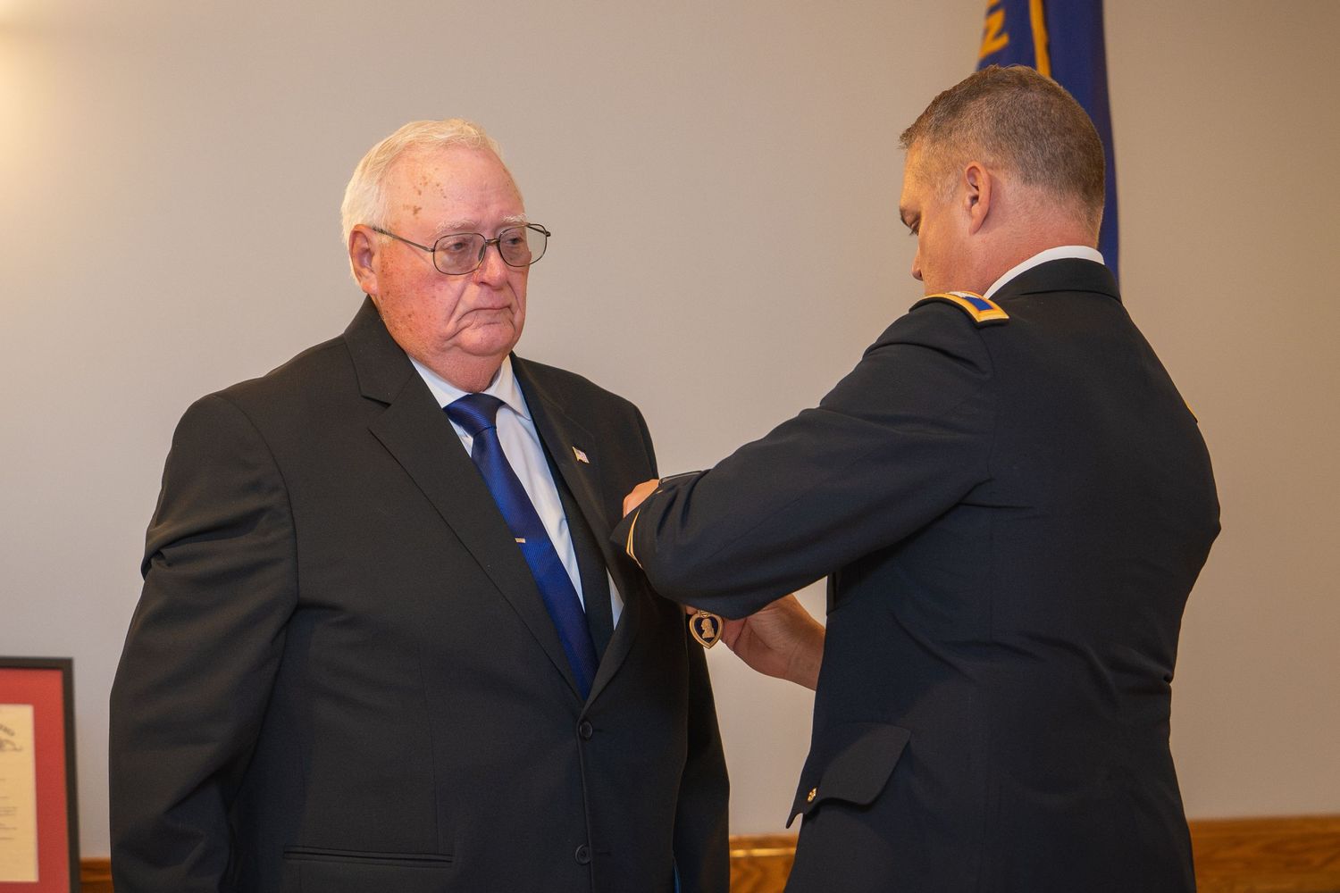 Military officer pins a medal on an older man in a suit during a formal award ceremony indoors.
