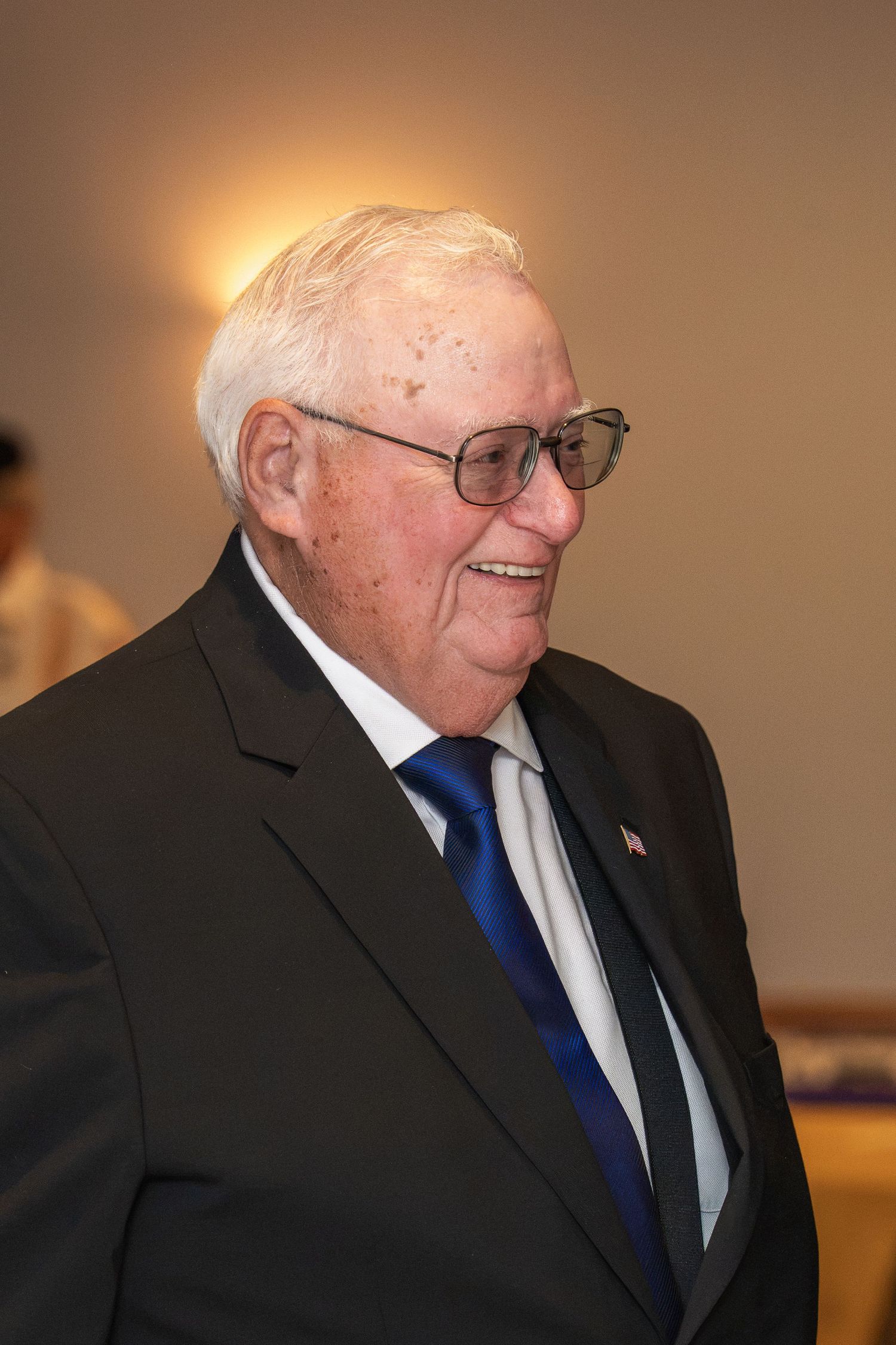 Elderly man in dark suit and blue tie wearing glasses, smiling warmly at a formal indoor event.
