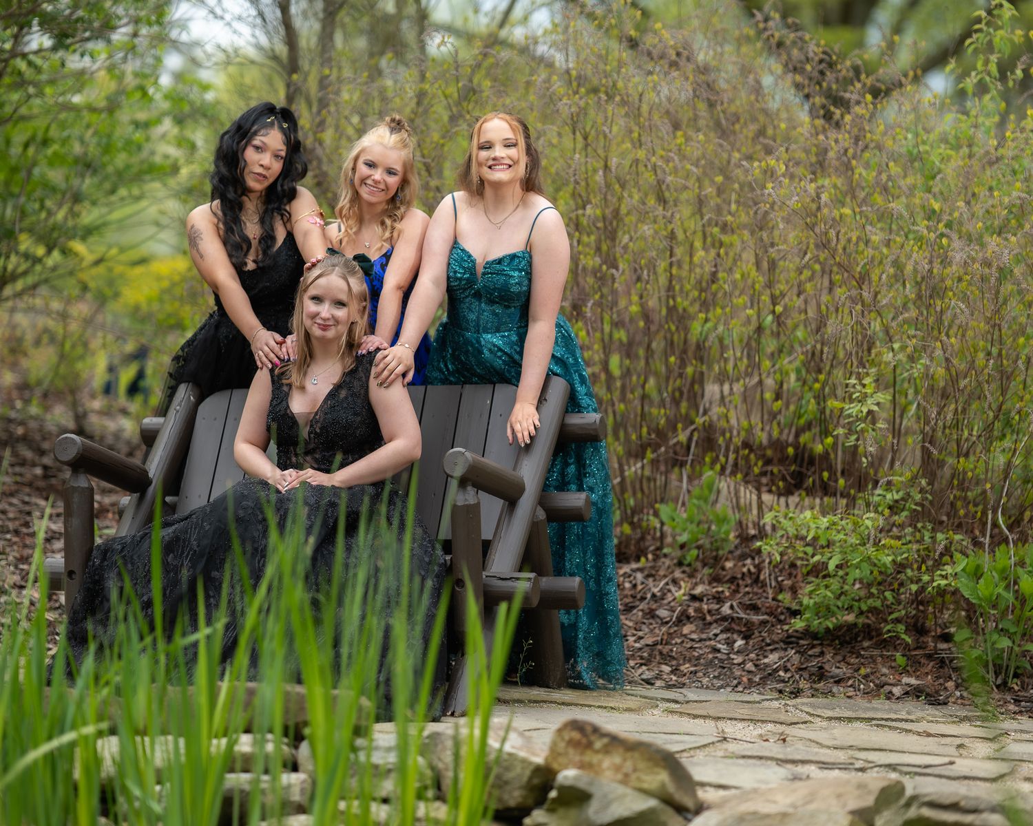 Four young women in elegant formal gowns pose together on a garden bench surrounded by lush greenery.