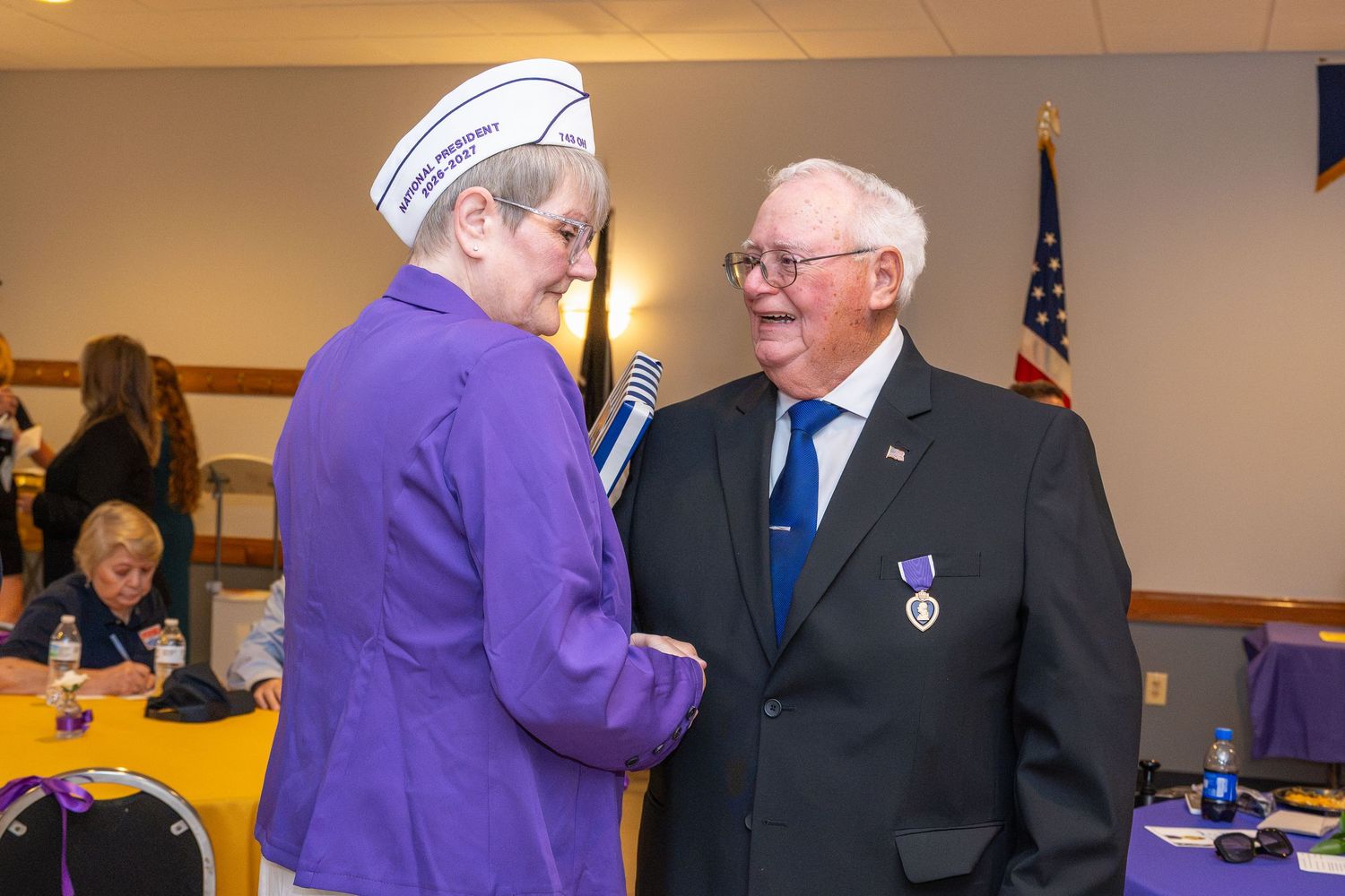 Two people in formal attire conversing at a purple and gold decorated indoor event.