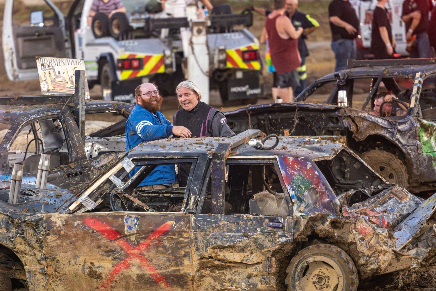 Two men smiling in a heavily damaged, mud-covered demolition derby car at an outdoor event.
