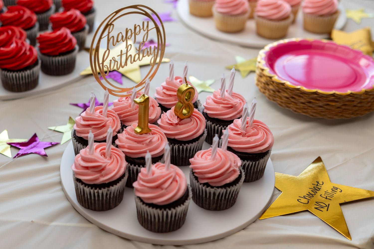 Chocolate cupcakes with pink frosting and a happy birthday topper on a white cake stand at a birthday party.