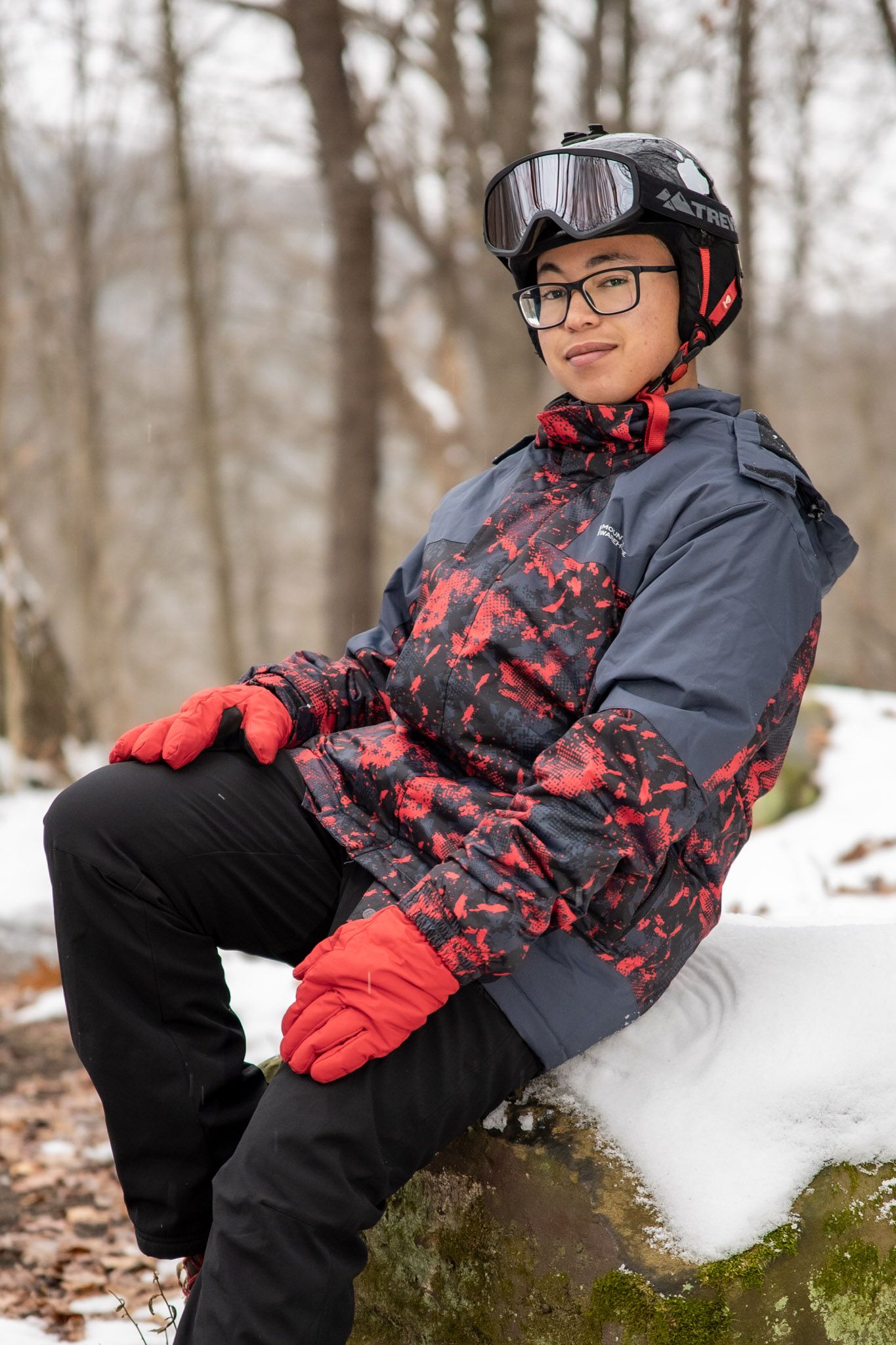 A person in winter gear sits on snowy ground wearing red and gray patterned outerwear.