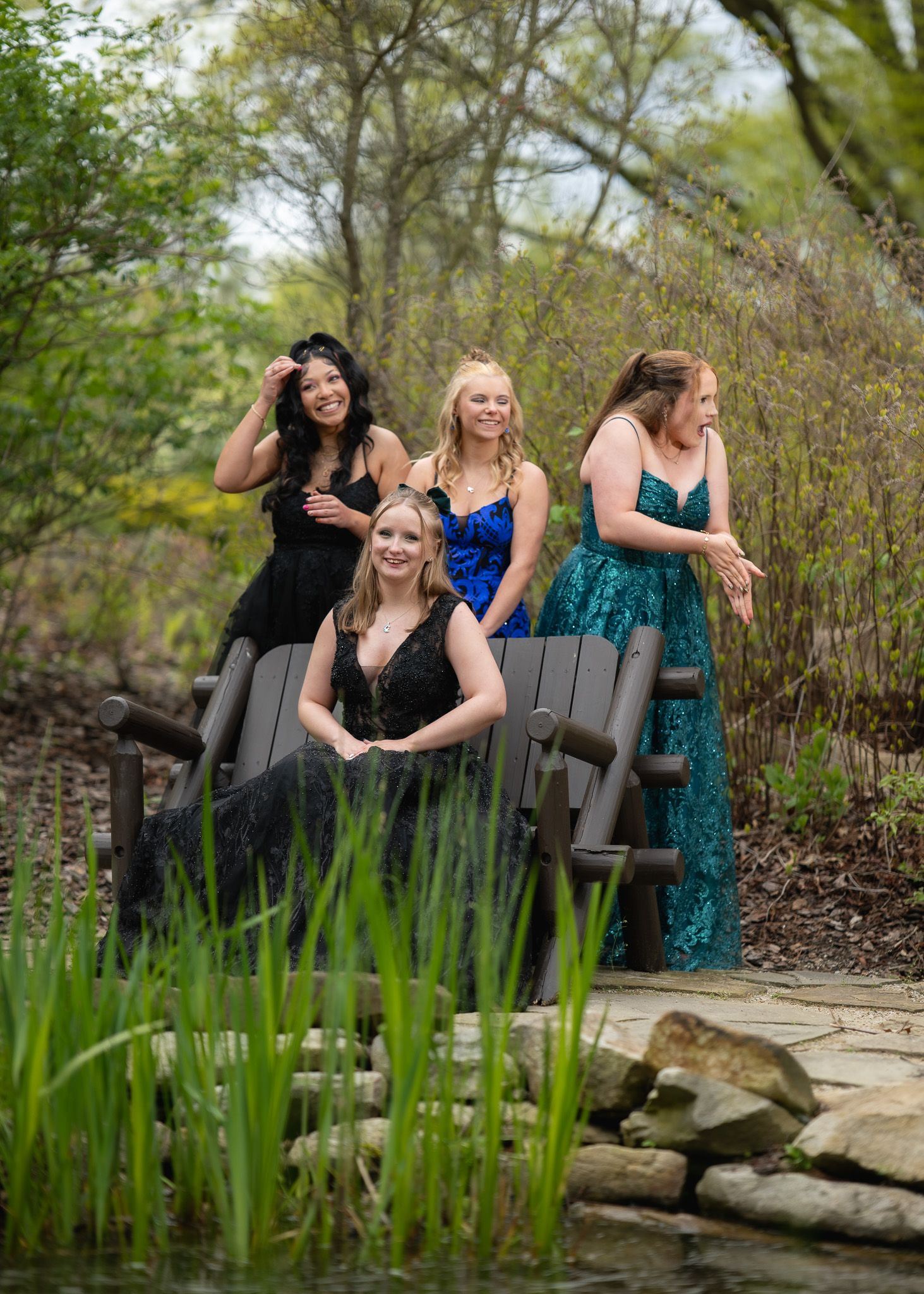 Four young women in formal gowns pose outdoors near a pond surrounded by lush greenery for prom photos.